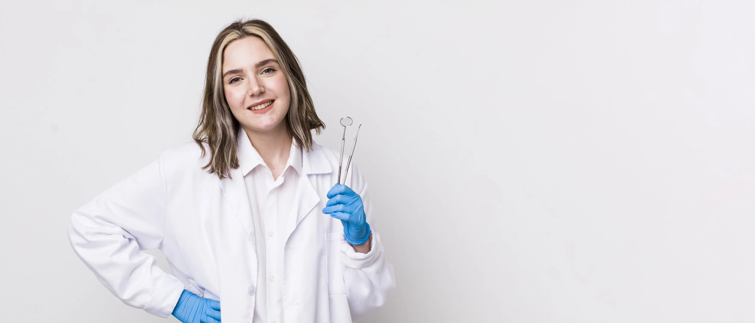 A young woman dentist wearing a white lab coat and blue gloves, holding medical tools, standing against a plain white background.