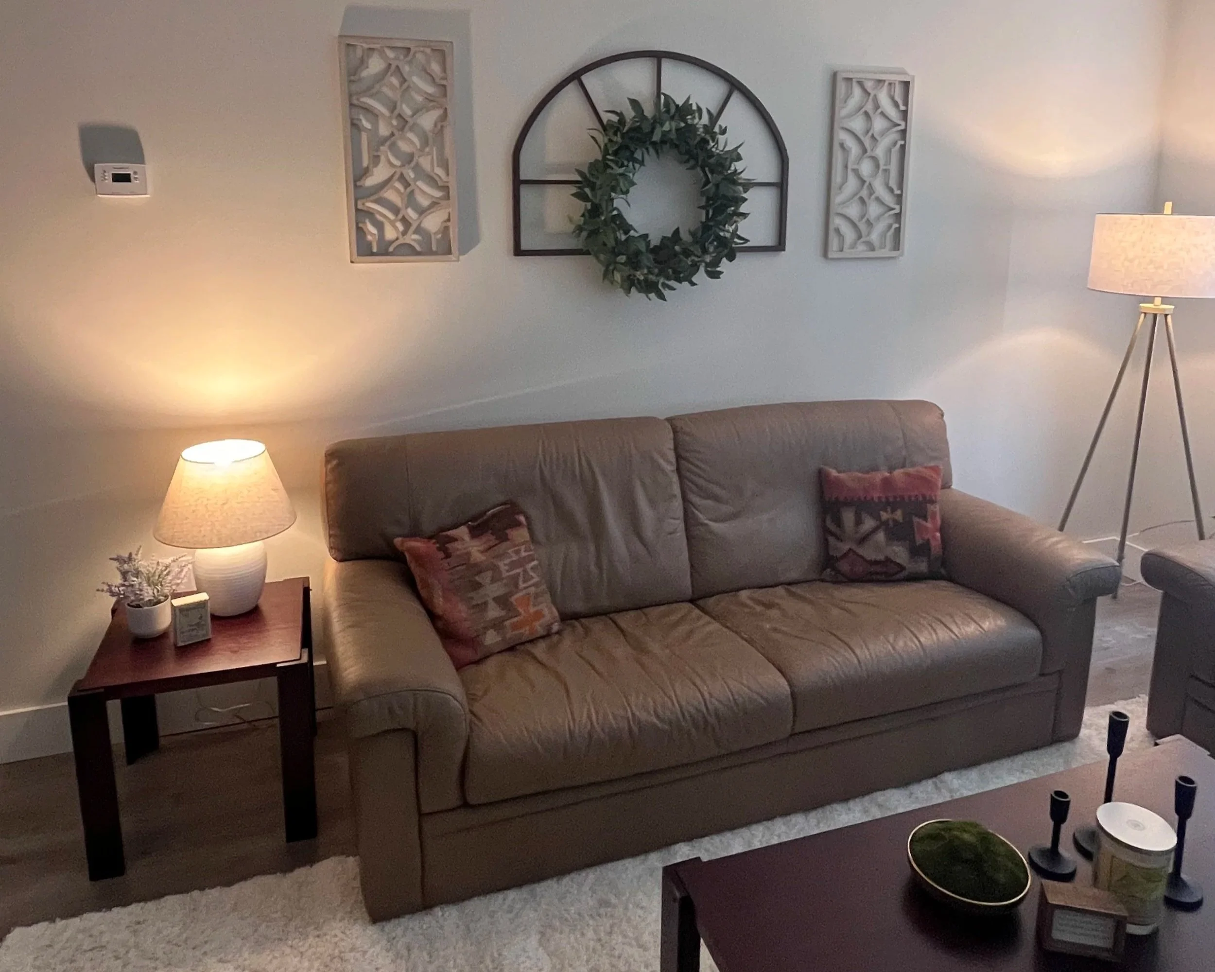 Living room with beige leather sofa, side table with lamp and small plant, wall decor including a wreath and decorative panels, using warm lighting.