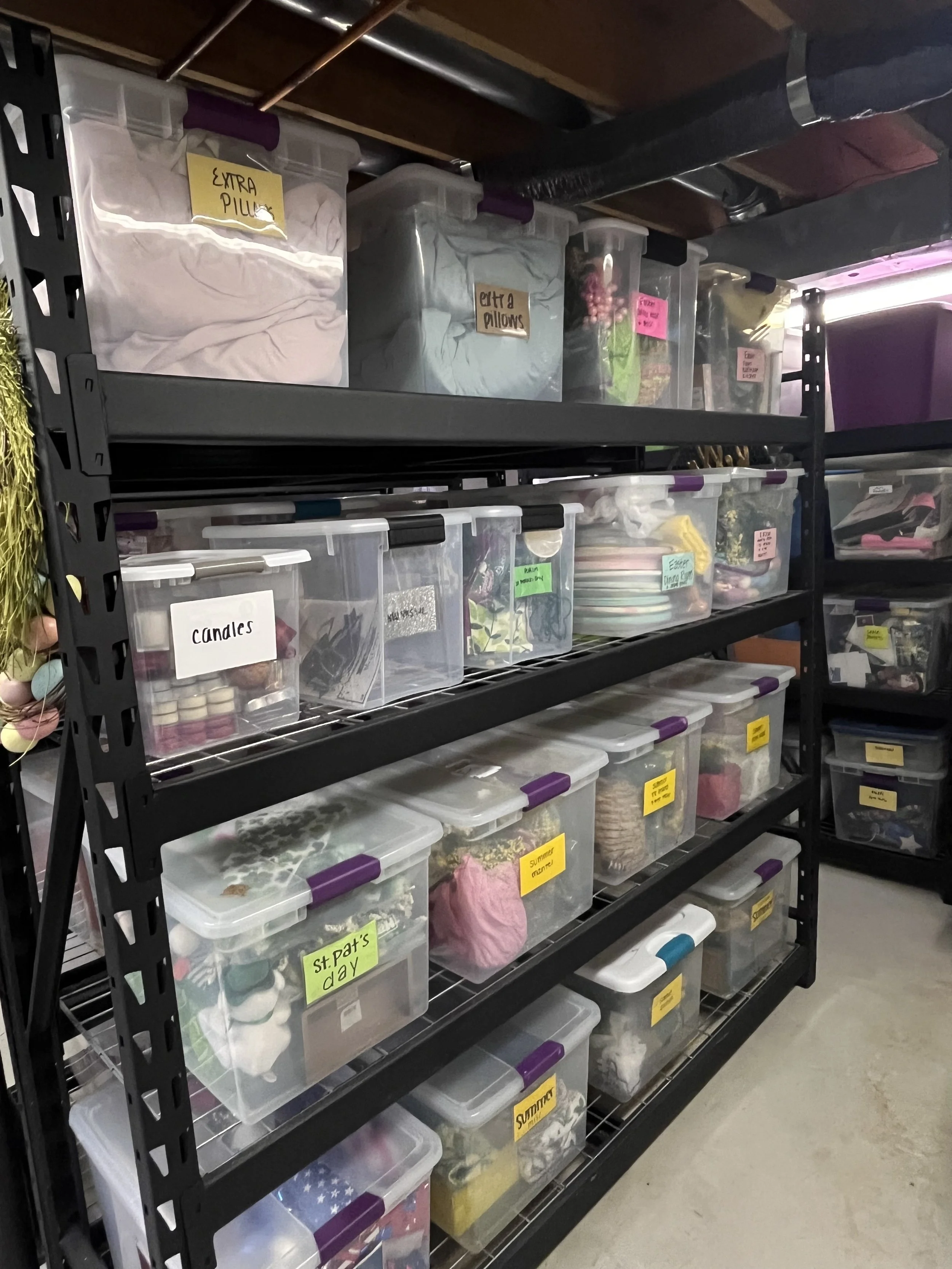 A black metal shelving unit filled with clear plastic storage bins labeled with various categories, containing clothing, decorations, and craft supplies, in a basement or storage room.