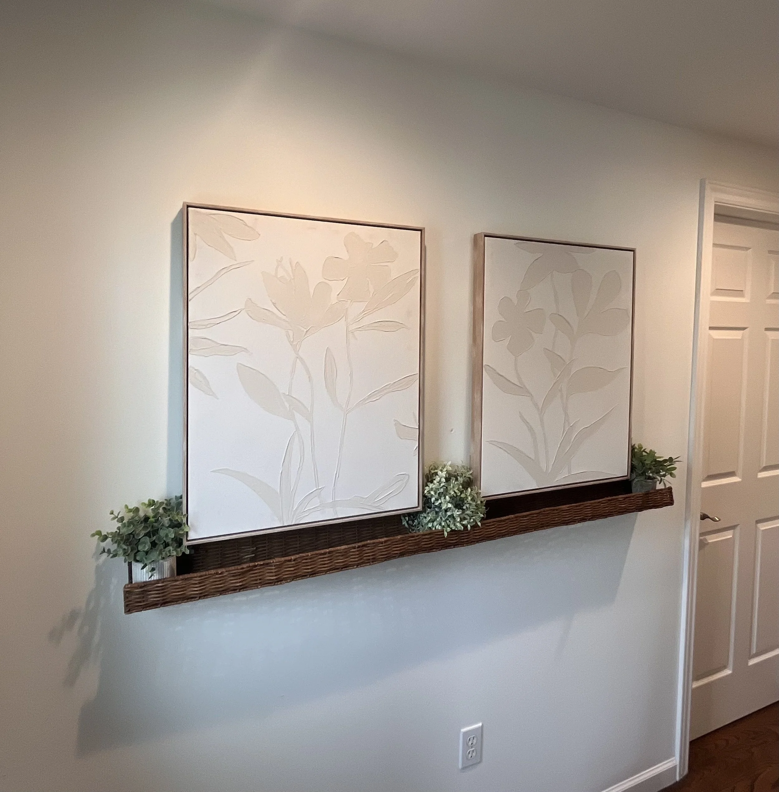 Wall decor with two framed floral prints, a wooden shelf with small potted plants underneath, and a nearby door.