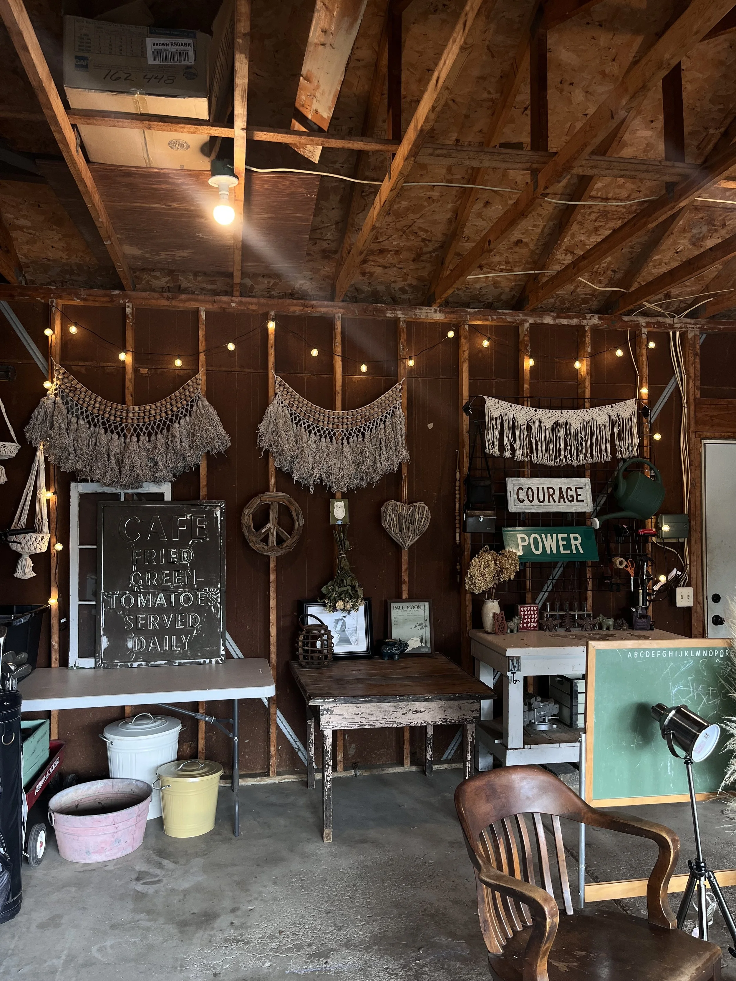 Interior of a rustic decorated room with wooden walls and ceiling, string lights, macrame wall hangings, a chalkboard menu, and various decorative items including peace, courage, and power signs, framed pictures, a chalkboard, and potted plants.