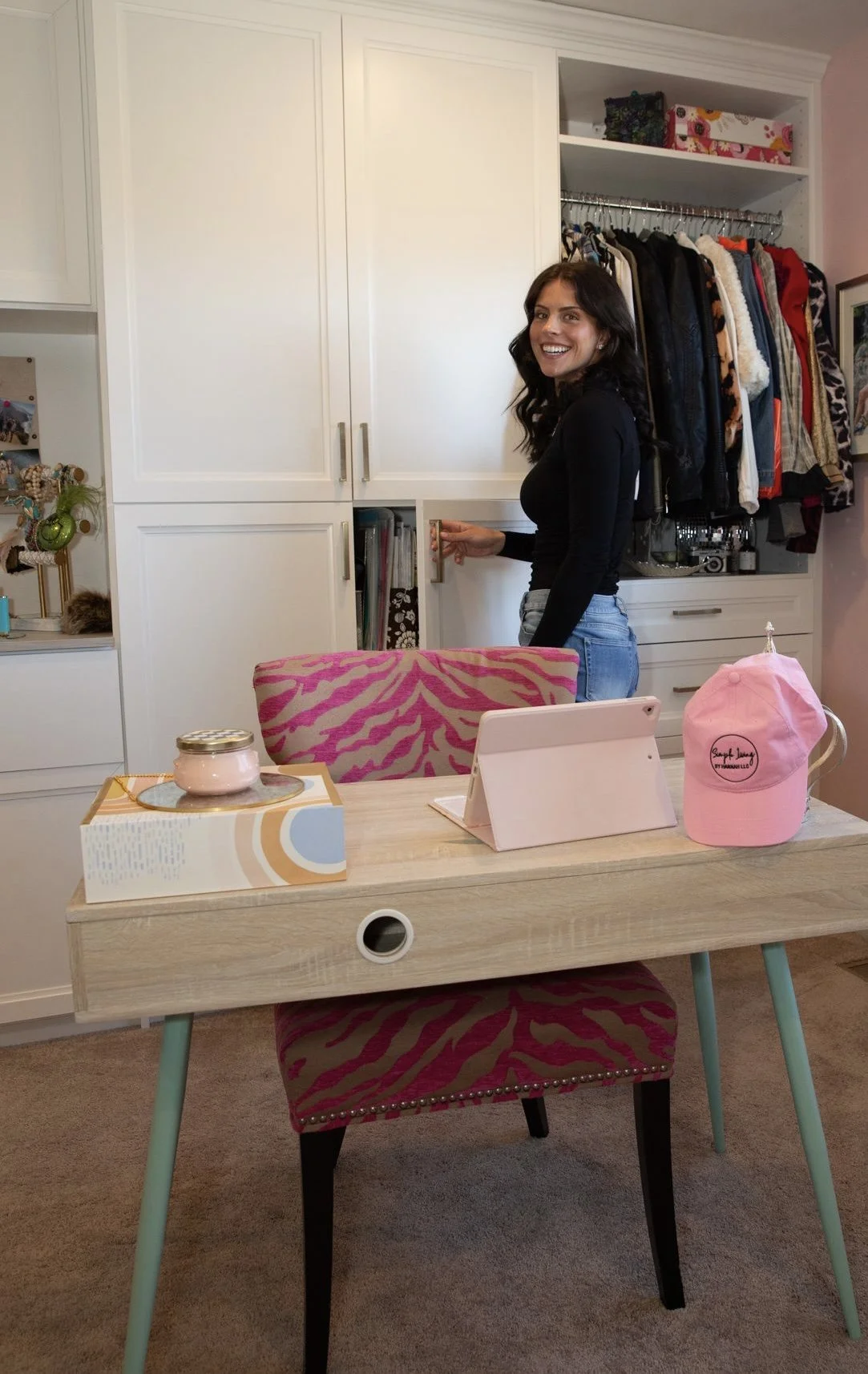 A woman with dark wavy hair wearing a black top and jeans standing near a closet with open white doors, smiling. In front made of light wood, a desk with a pink and red zebra-patterned chair, holding a pink baseball cap, a tablet, a candle, and a notepad.