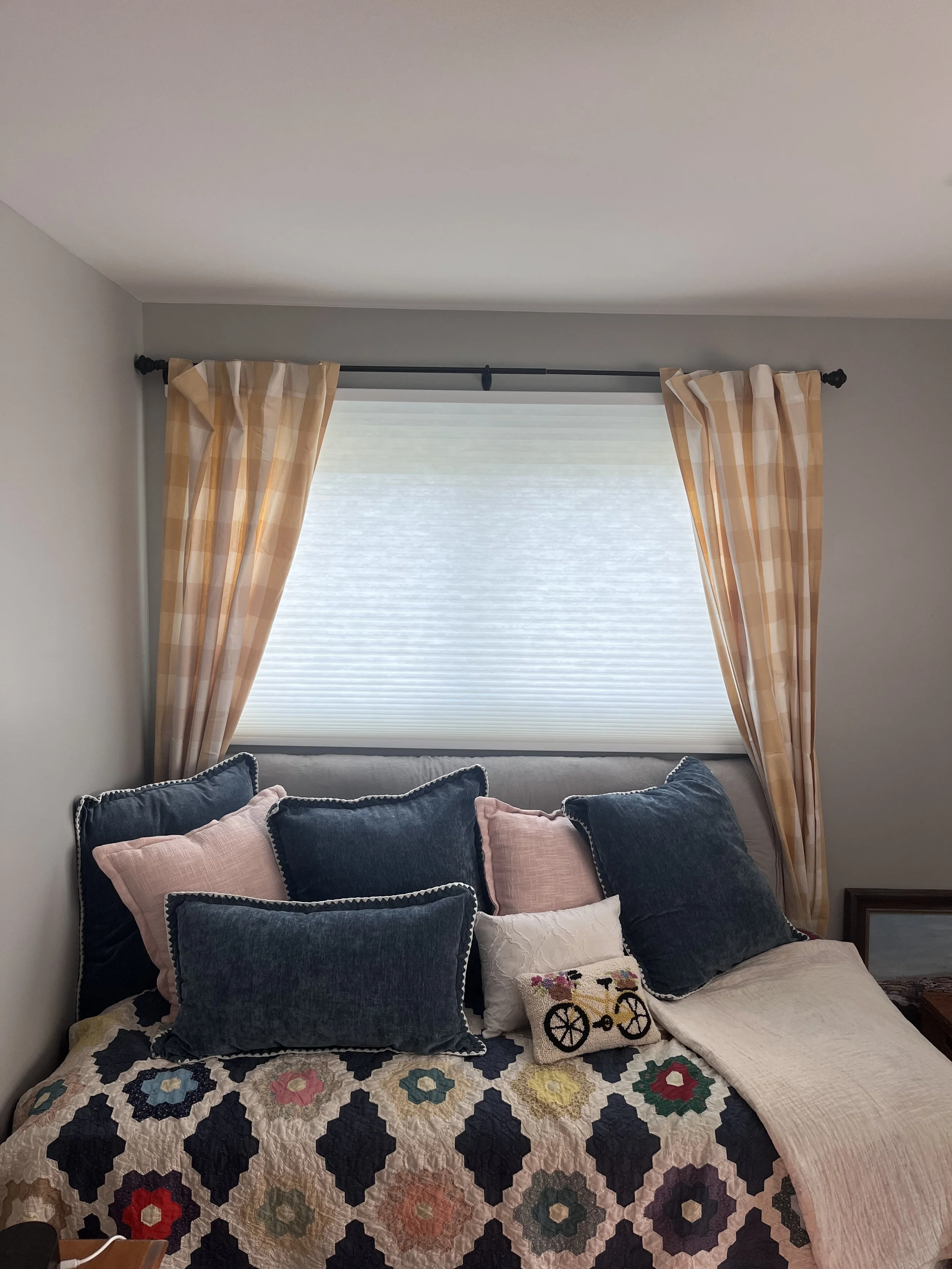 A bedroom with a window covered by beige checkered curtains and a white pleated blind. The bed has a quilt with a floral pattern and is decorated with pillows in pink, dark blue, and a decorative pillow with a bicycle embroidered on it.