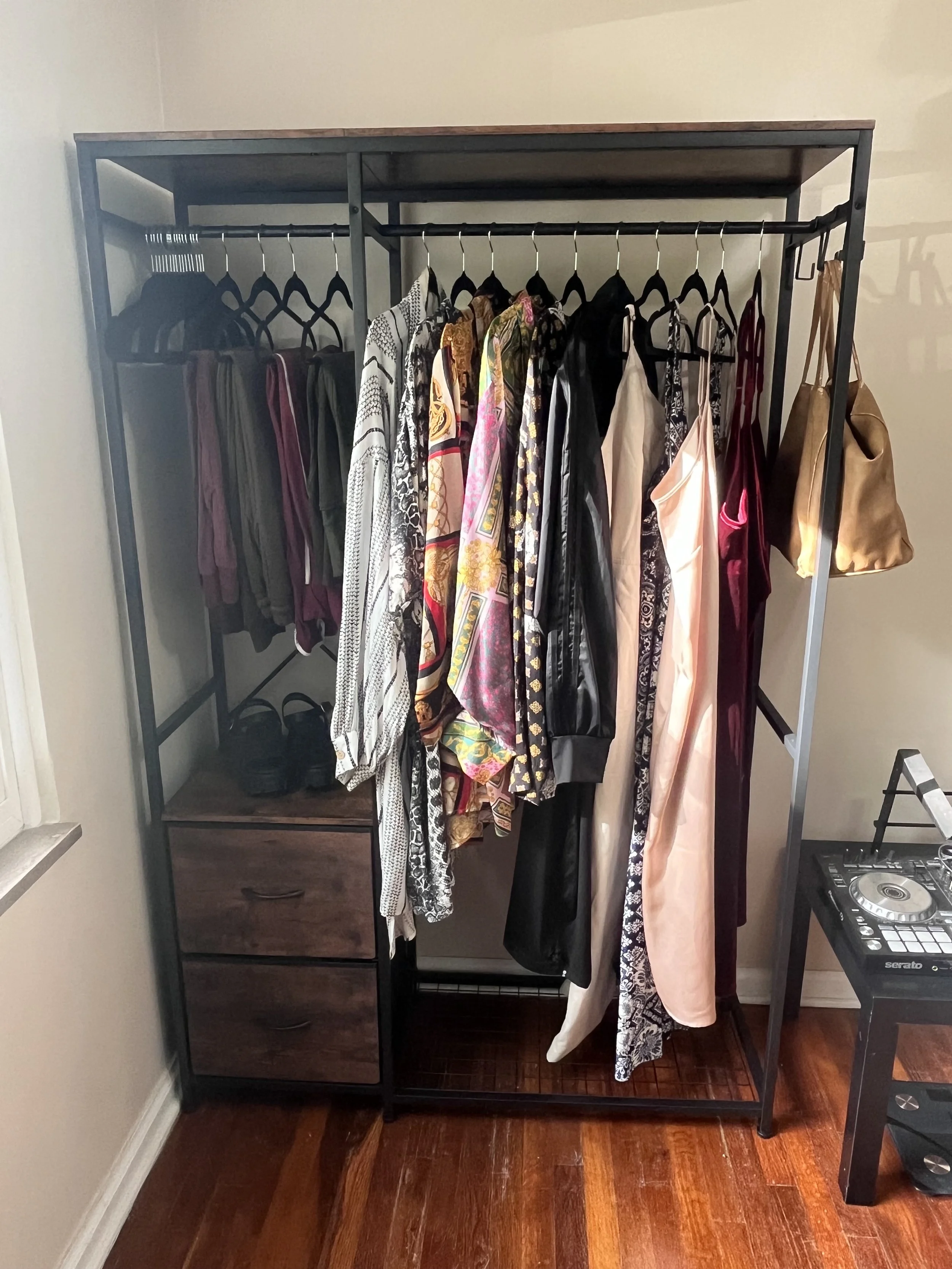 A black metal clothing rack with a wooden top, containing various hanging clothes including patterned, black, and light-colored garments. There are two drawers at the bottom left, and two black shoes on the middle shelf. A tan handbag hangs on the ri