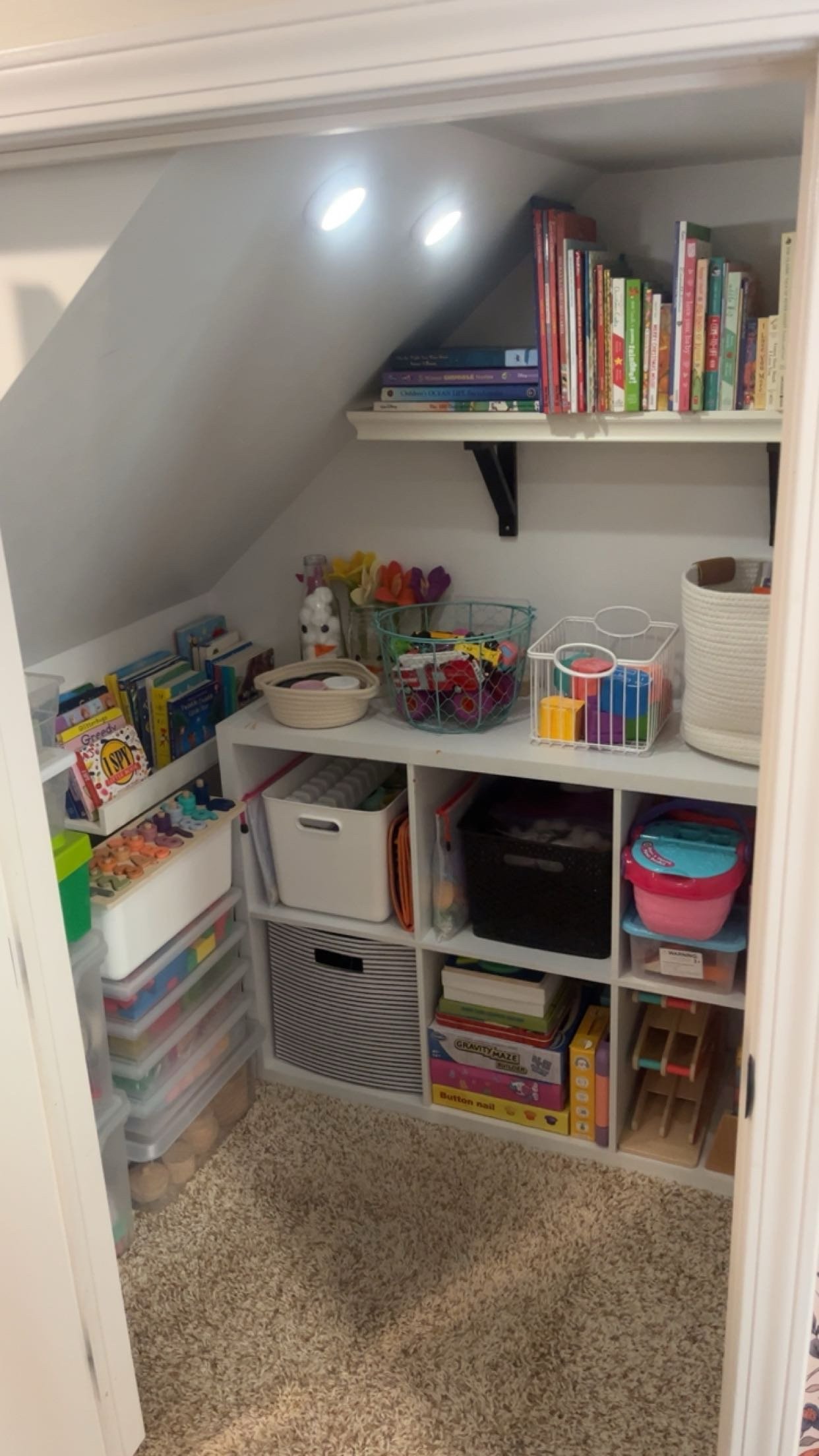 Organized toy and book storage area in a room with a sloped ceiling, including shelves filled with books, toys, and storage bins.