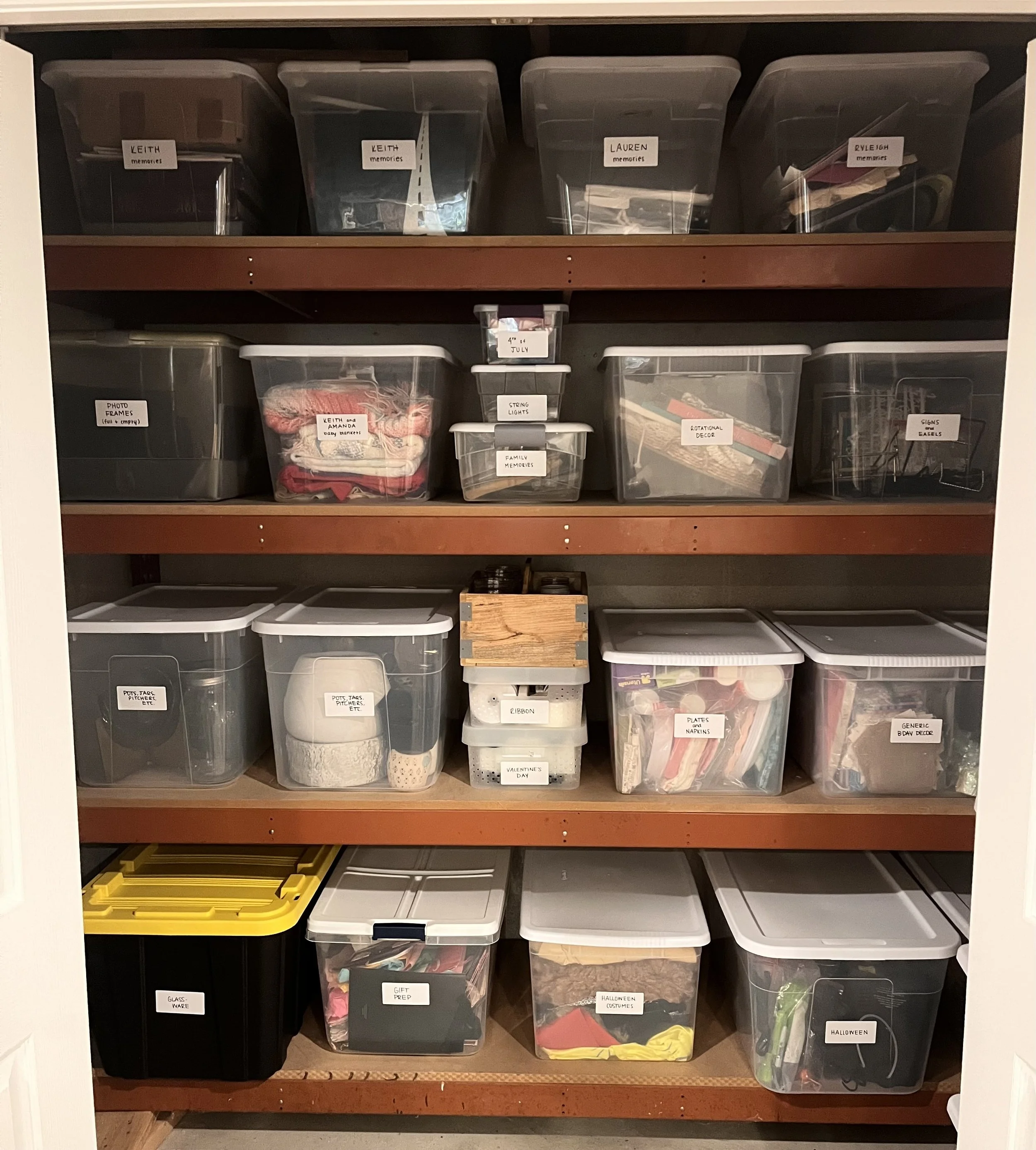 A wooden shelf with multiple clear plastic storage bins labeled with various categories such as memories, decorations, tags, and holiday items like Halloween costumes and Christmas gift prep.
