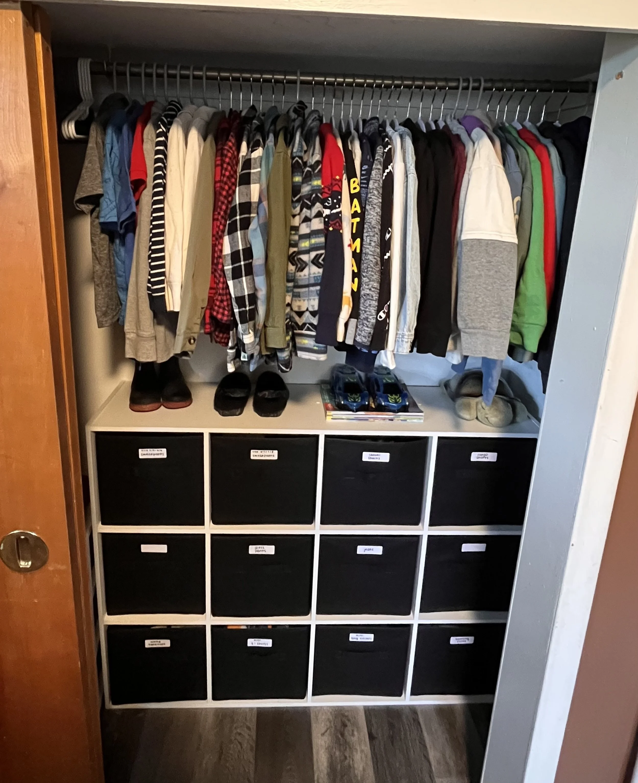 Open closet with hanging clothes on a rod above black storage bins labeled for different clothing categories.