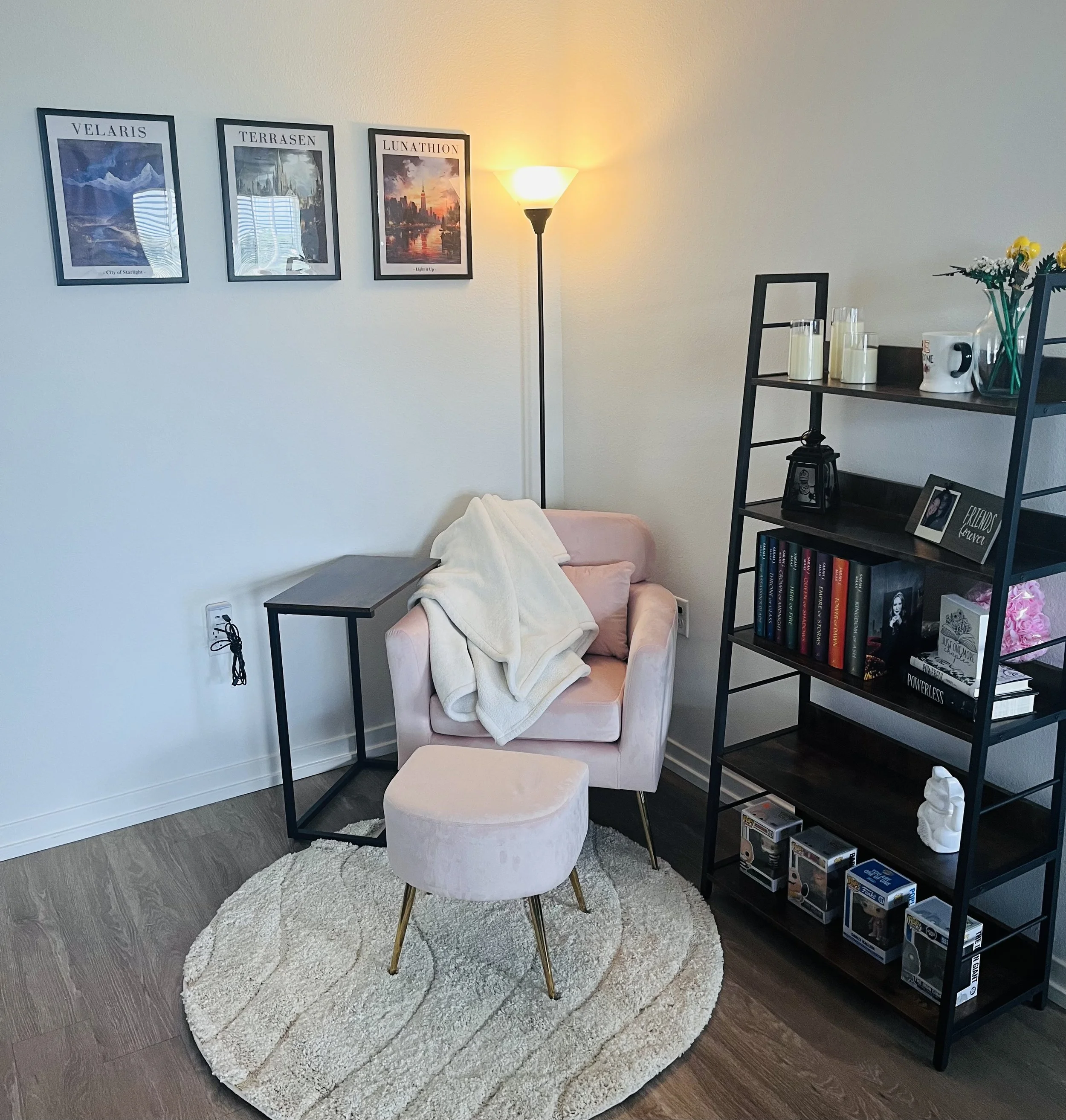A cozy corner of a living room with a pink armchair, small pink ottoman, and a cream-colored blanket draped over the arm. A black metal side table is beside the armchair. A round, cream-colored rug is beneath the furniture. A bookshelf with books, ph