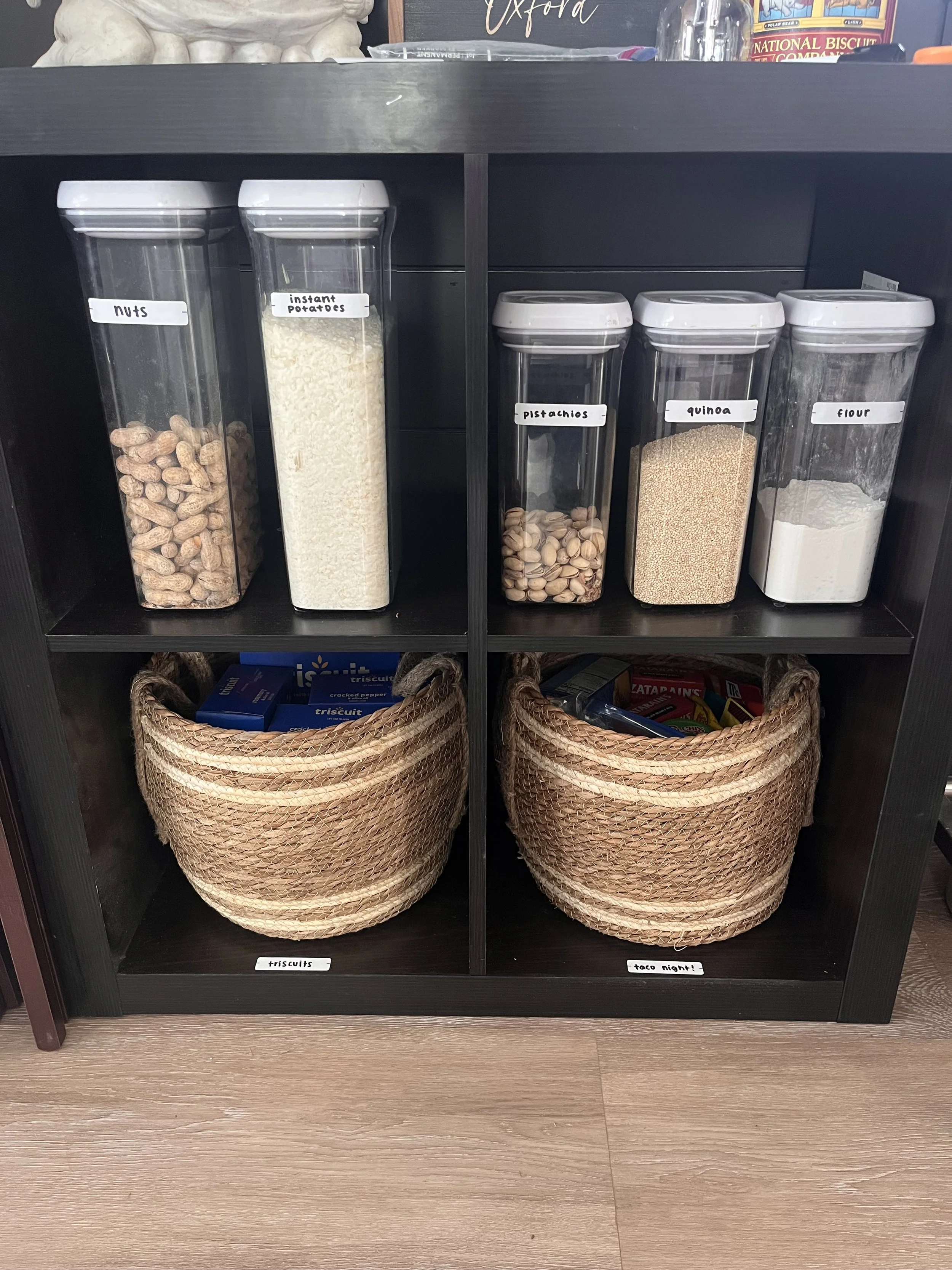 Shelf with containers of nuts, instant potatoes, pistachios, quinoa, and flour on top. Wicker baskets labeled 'triscuits' and 'taco night' on the bottom shelf.