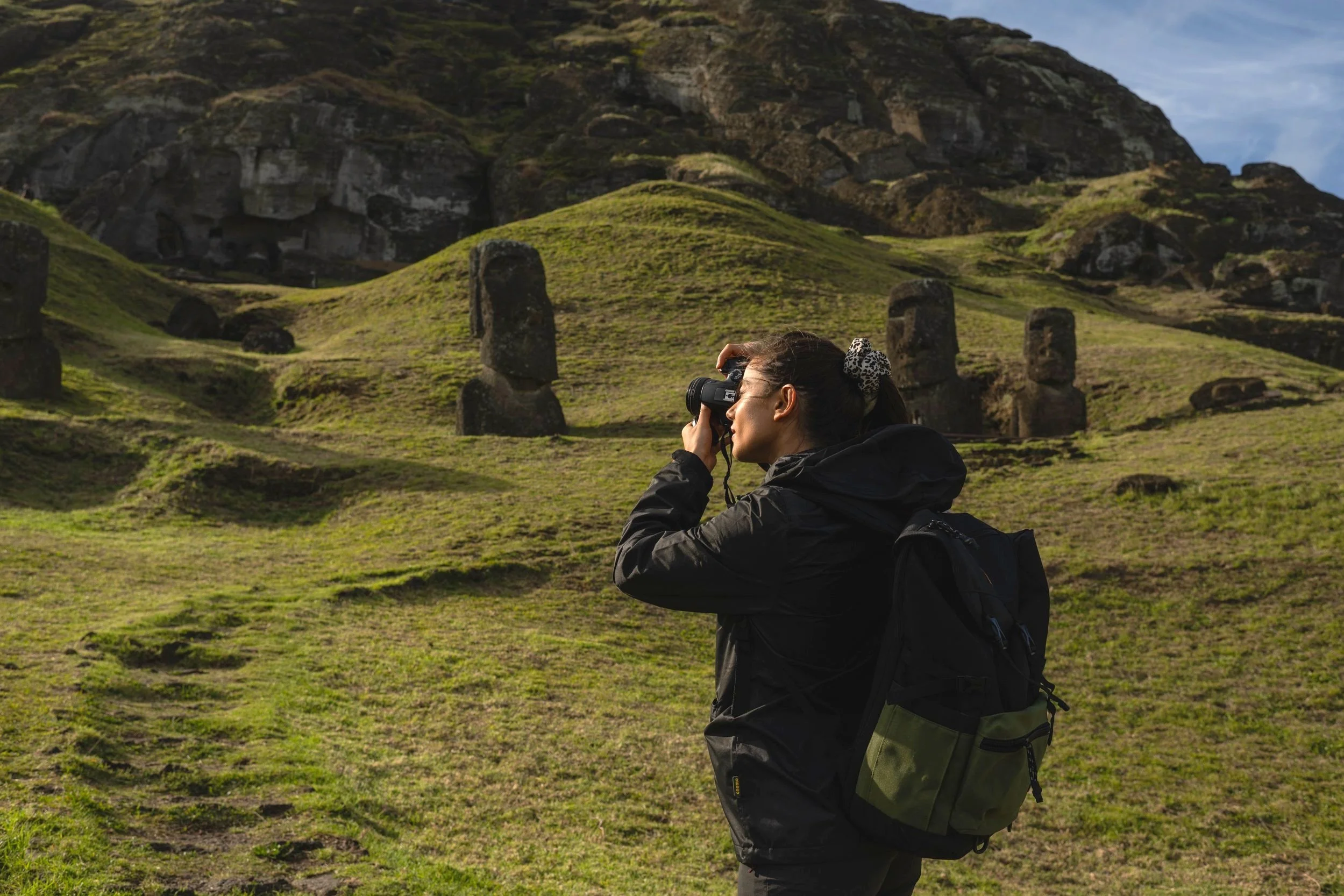 Persona tomando fotos a estatuas moái en Isla de Pascua, terreno verde y colina de fondo.