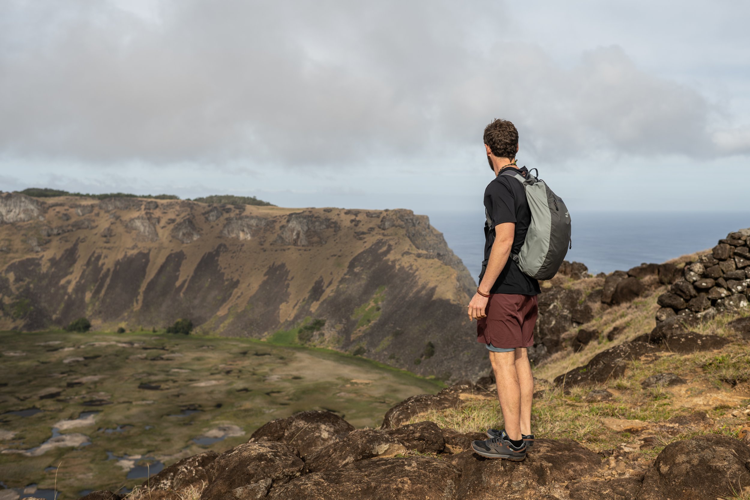 Hombre con mochila mirando un paisaje montañoso con el mar al fondo.