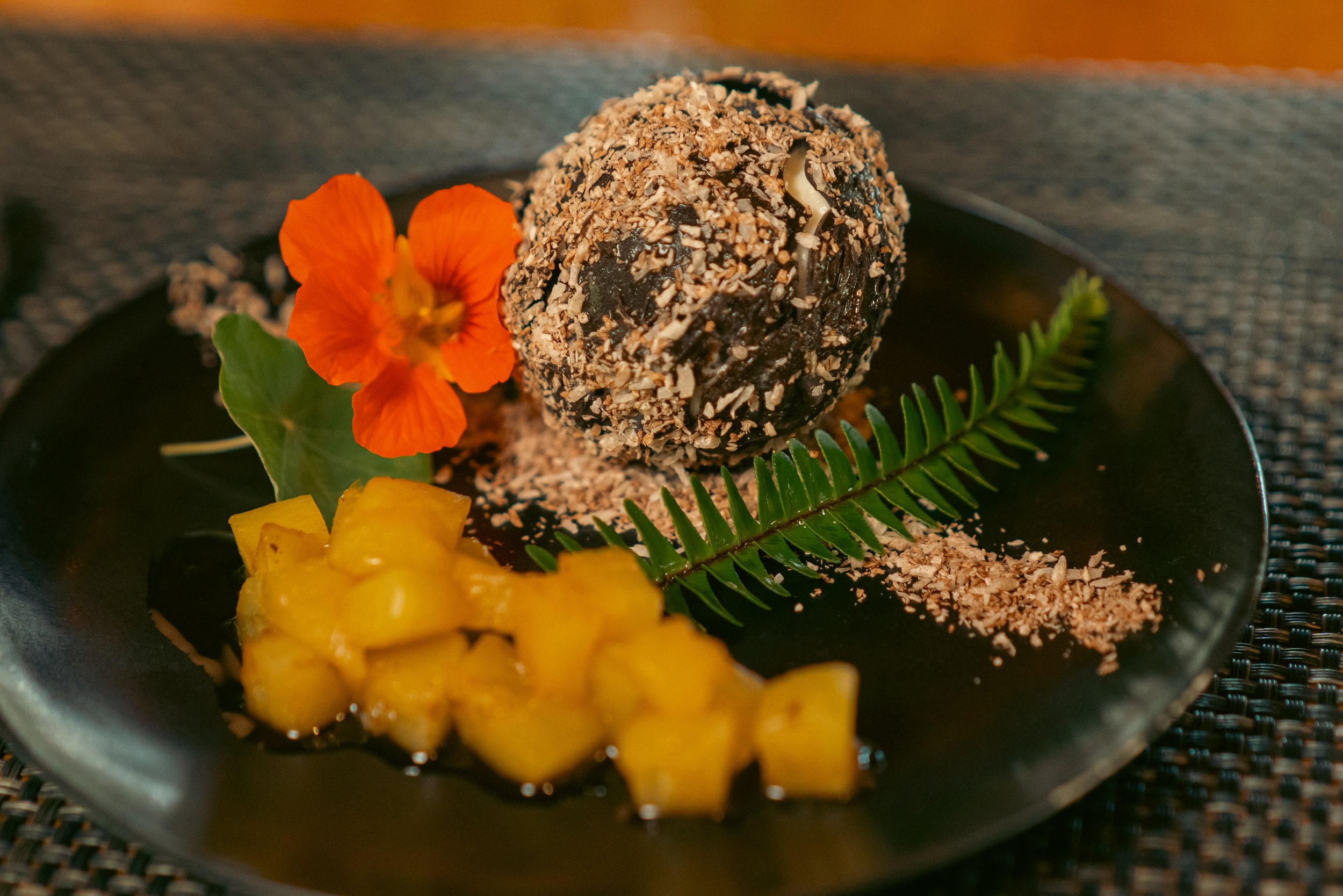 Plato decorativo con bola de chocolate espolvoreada, trozos de fruta amarilla y flor naranja, sobre hoja verde.