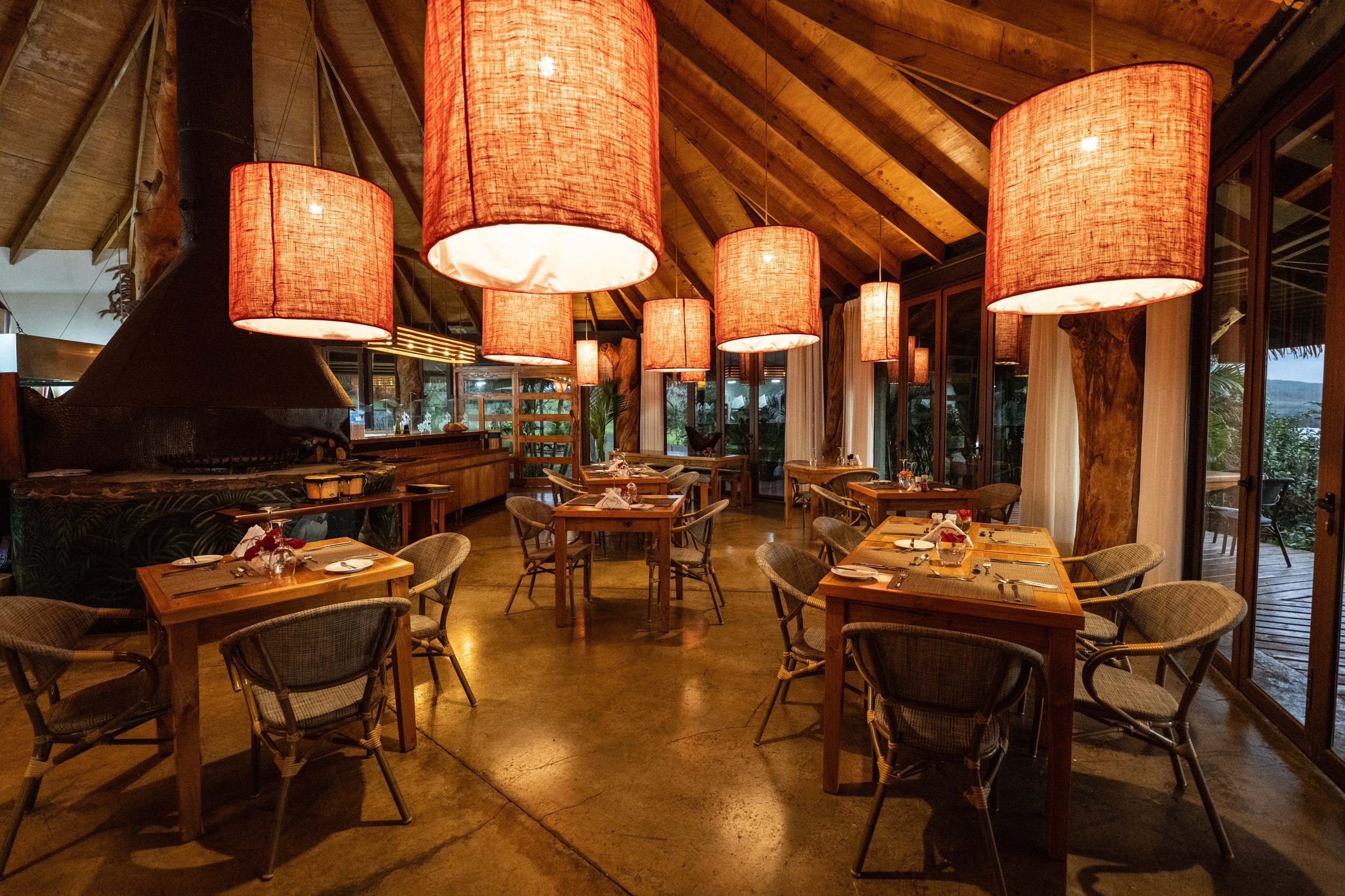 Interior de un restaurante acogedor con mesas de madera, sillas, grandes lámparas colgantes y una decoración rústica.