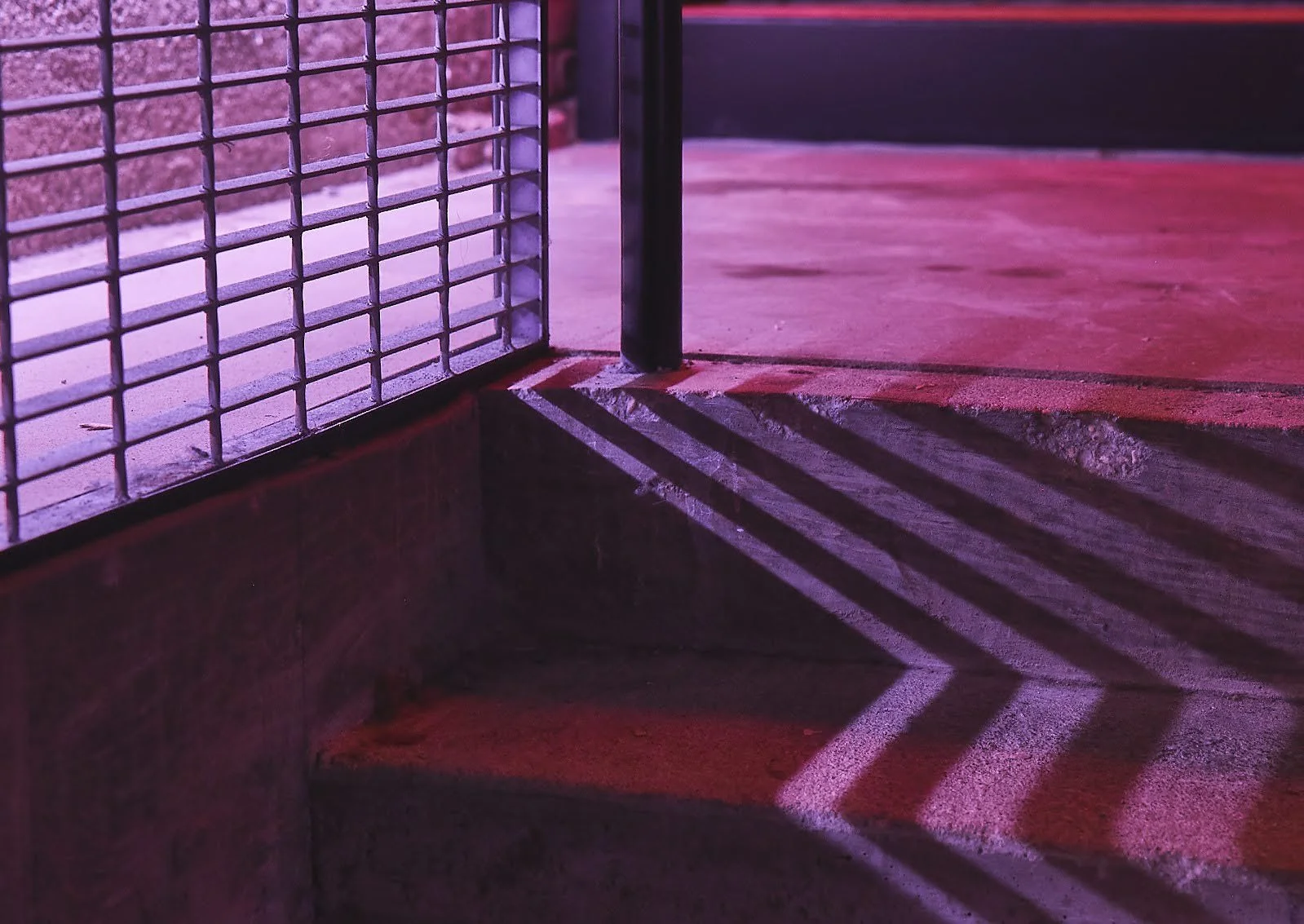 Shadow of a metal grid fence cast onto a concrete surface with purple and pink lighting.