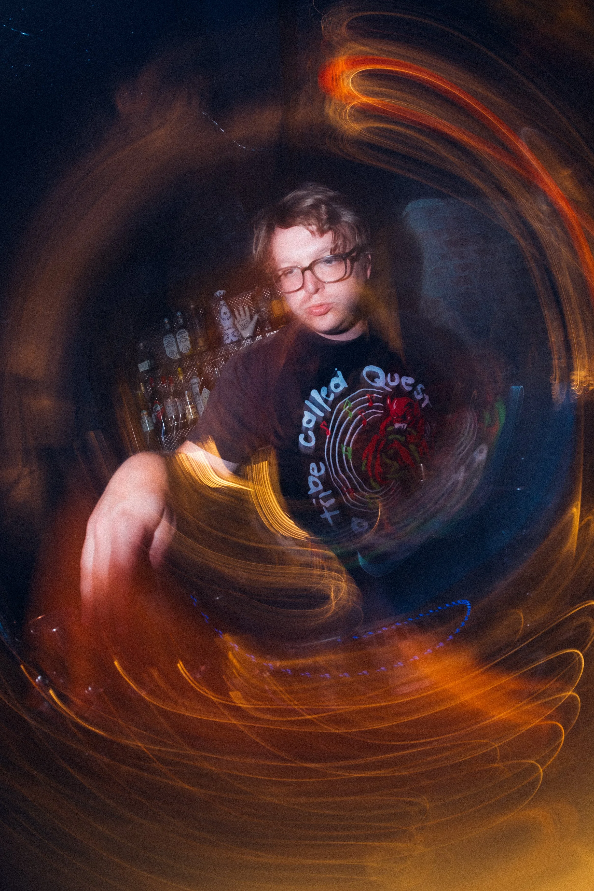 A person with glasses and a dark T-shirt standing in a dimly lit bar with swirling light effects around them.
