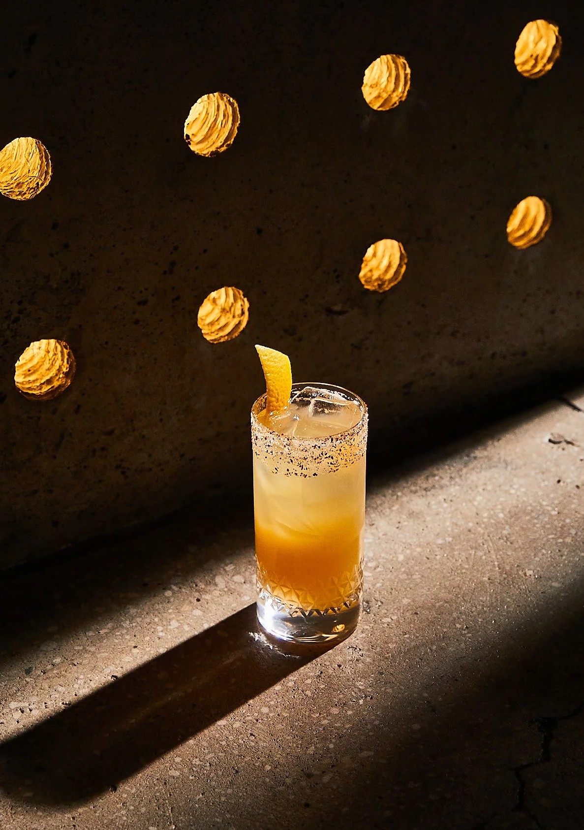 A tall glass of cocktail with a lemon wedge garnish, salt rim, and ice, casting a shadow on a textured surface. Multiple cookies are suspended in the dark background.