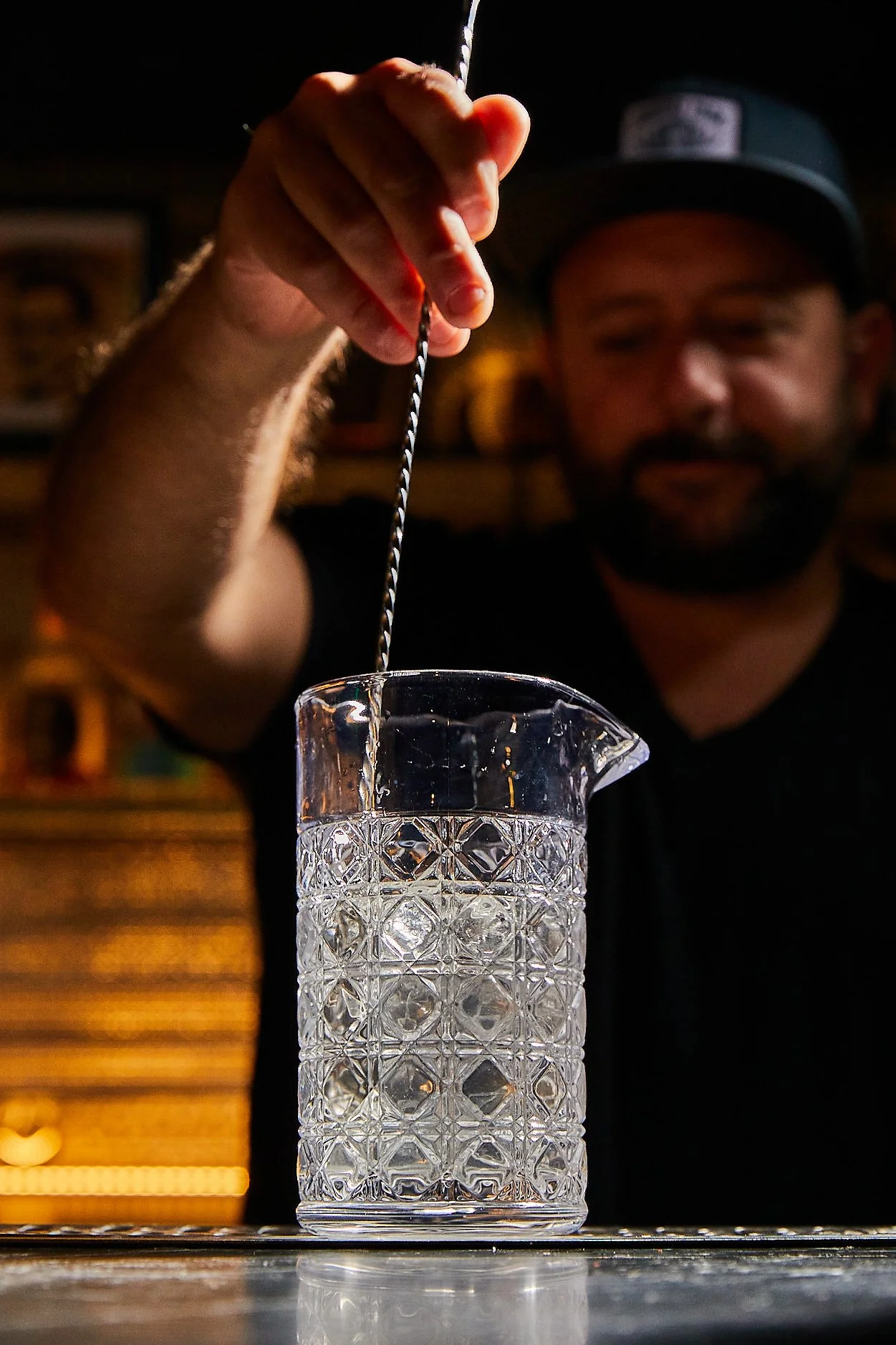 A man wearing a black shirt and baseball cap is stirring a liquid in a cut glass measuring pitcher with a strainer stick, in a dimly lit setting.