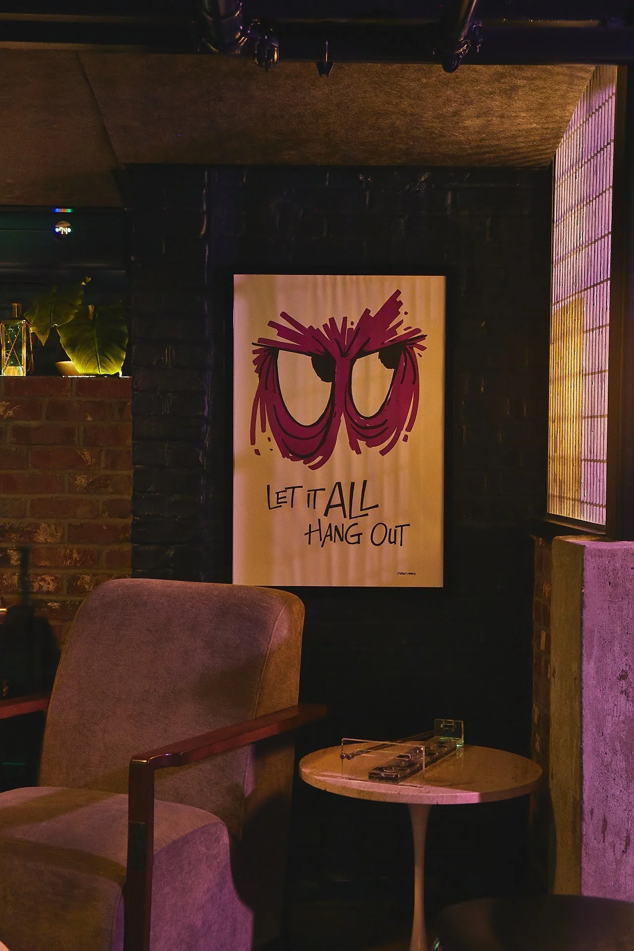 A framed cartoon-style artwork of purple animal eyes with bushy eyebrows and glasses, with the quote 'Let It All Hang Out,' hangs on a dark brick wall in a cozy, dimly-lit room.