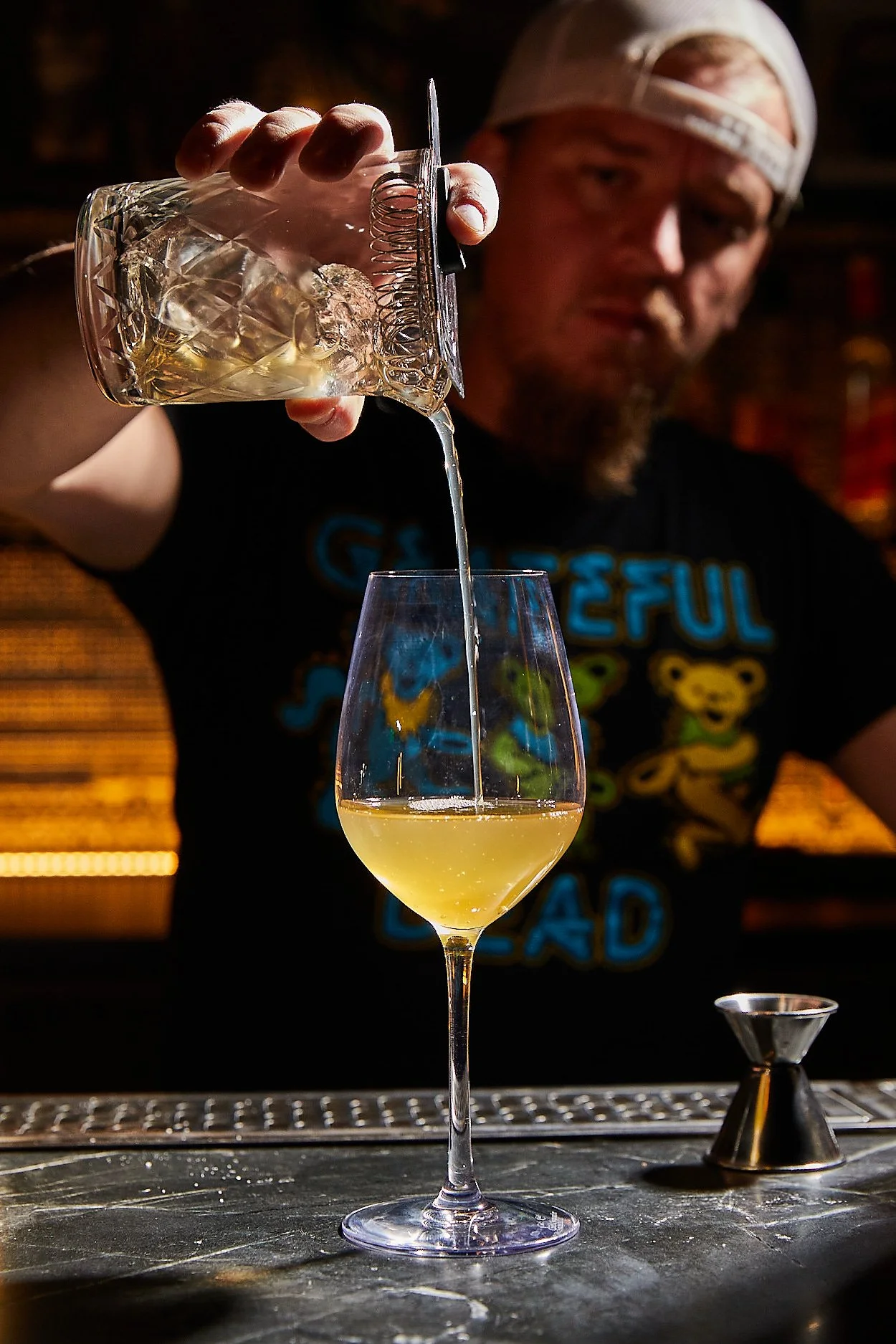 Bartender pouring a yellow cocktail into a wine glass with ice, with a cocktail shaker nearby.