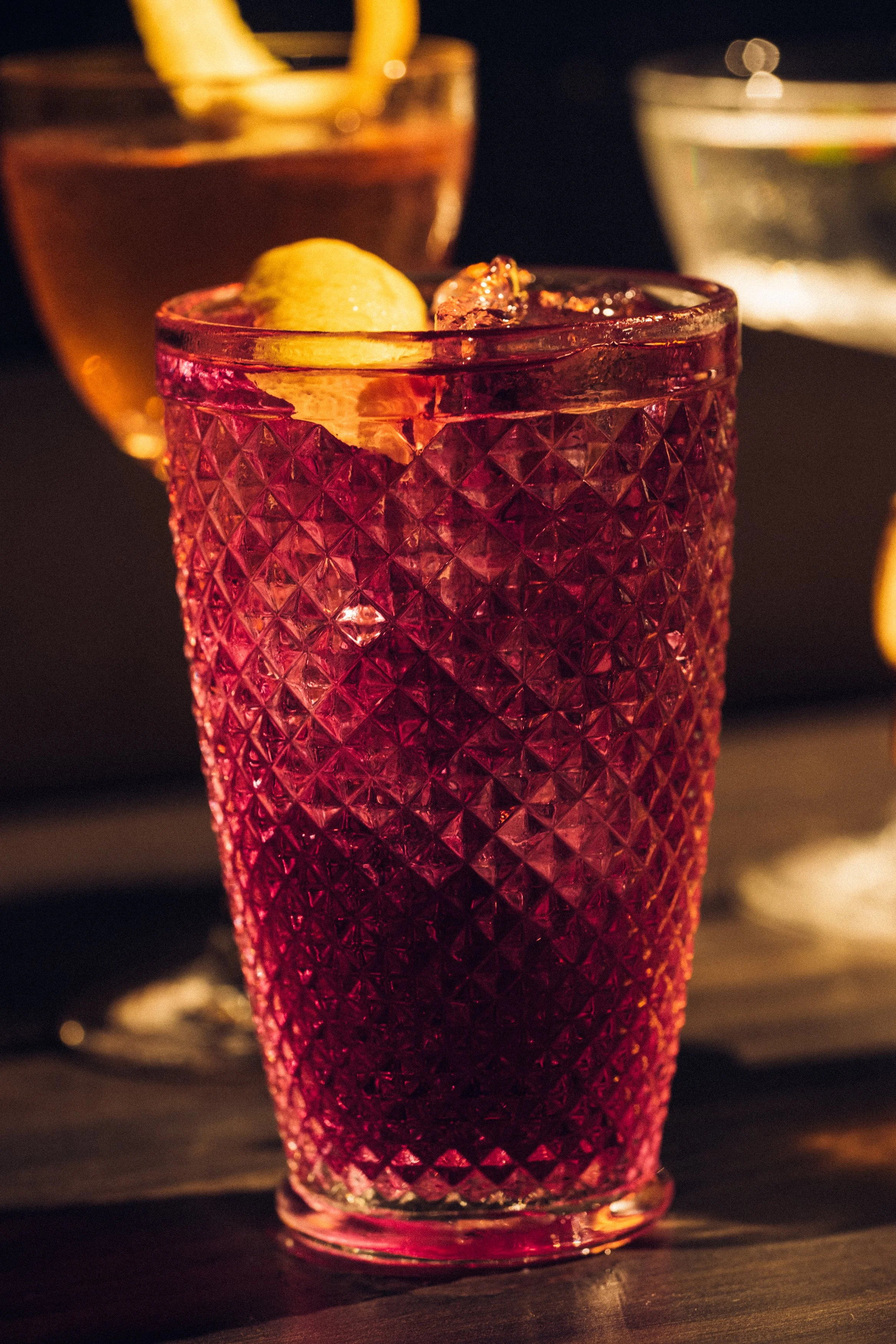A pink textured glass of a berry cocktail with a lemon slice and ice, with other drinks in the background.