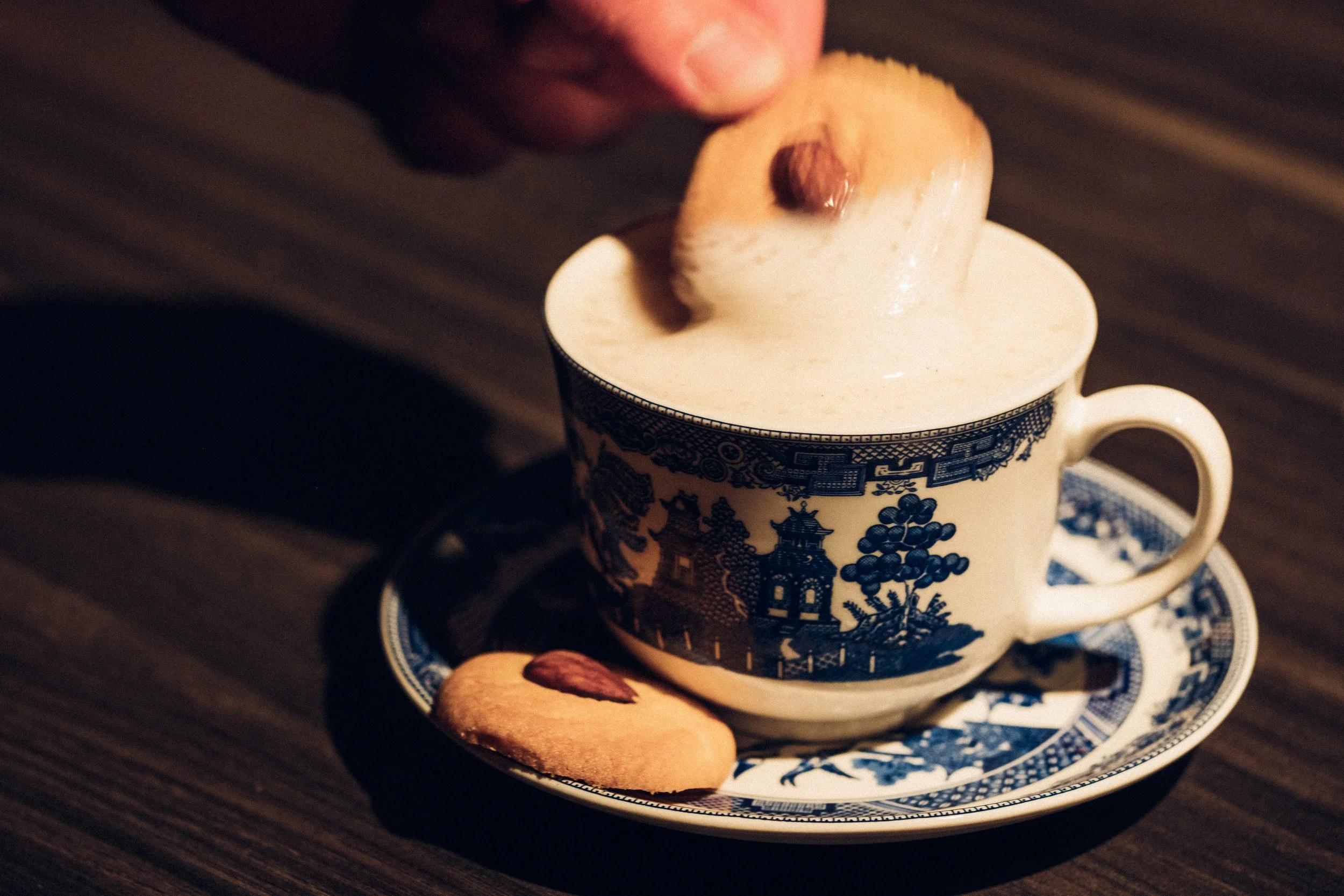 A hand holding a cookie over a cup of frothy coffee on a patterned saucer and cup with a scenic design, with an additional cookie on the saucer.