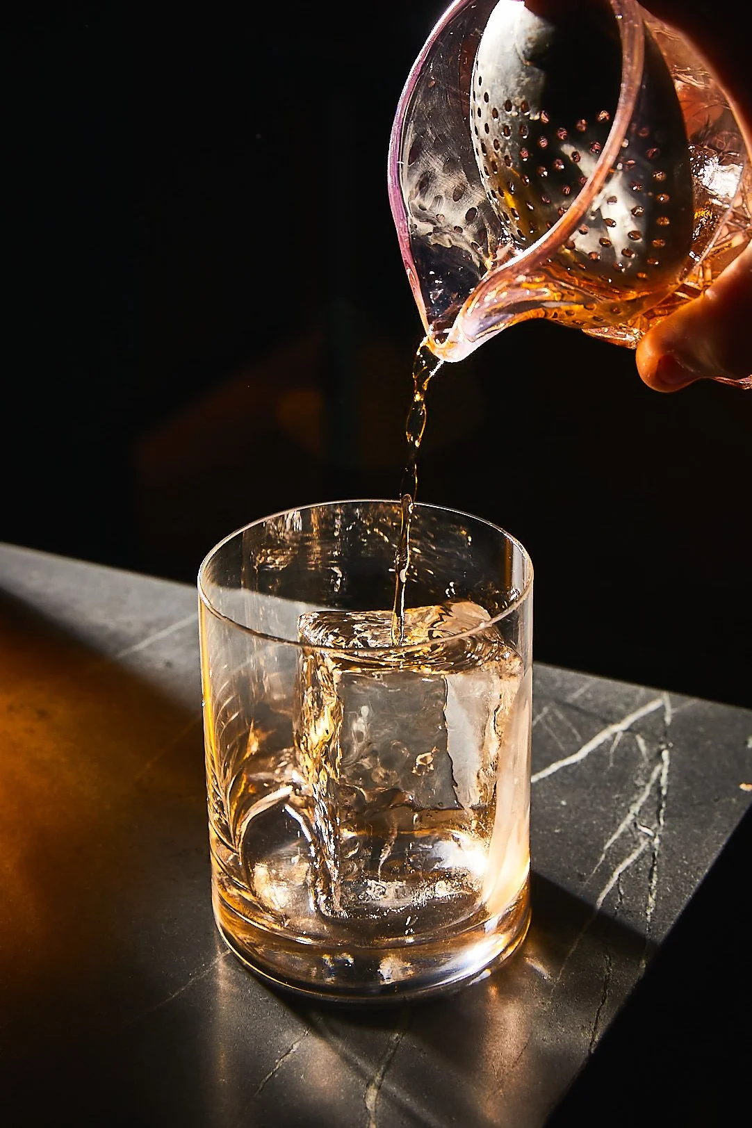 A hand pouring liquid from a glass pitcher into a glass with ice on a black marble surface.