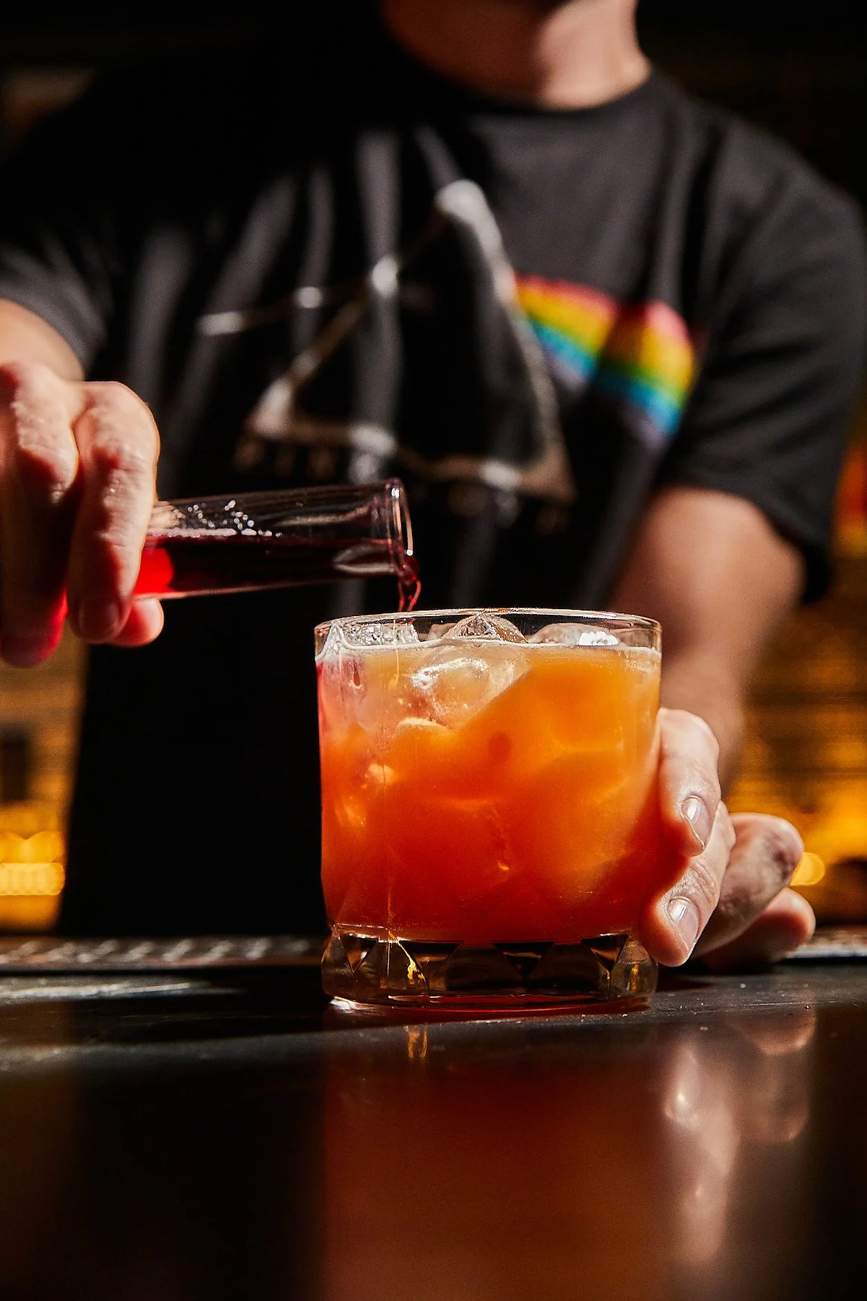 Person pouring red wine into a glass containing orange cocktail with ice on a bar counter.