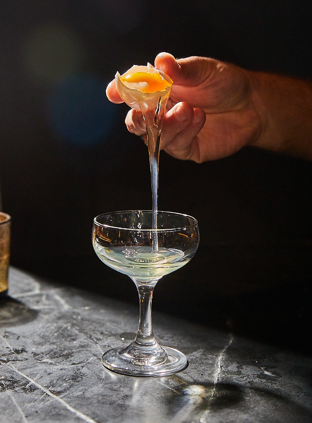 A hand cracking open a soft-boiled egg into a cocktail glass.