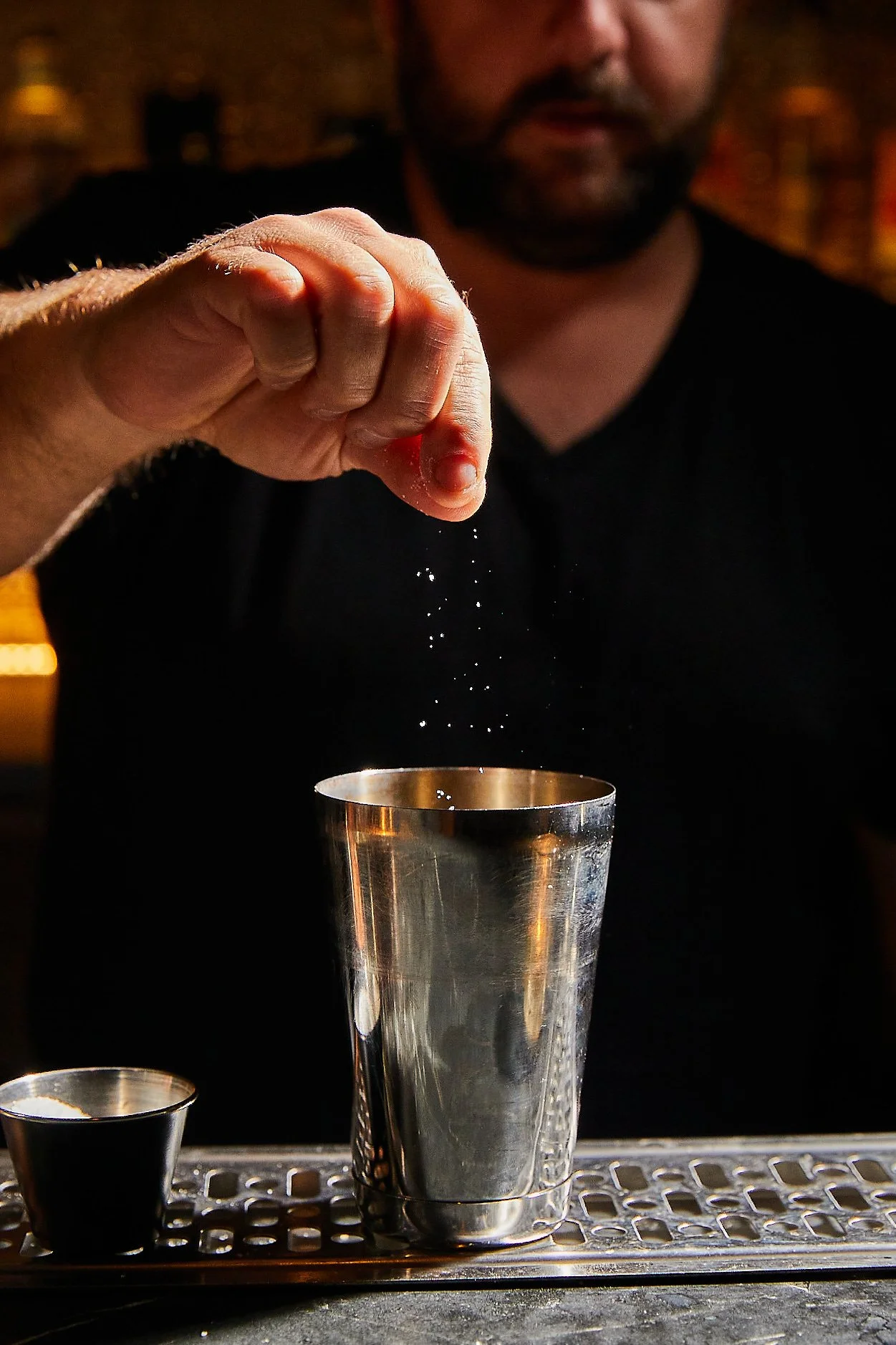 A bartender sprinkling salt into a metal shaker. The person's face is partially visible, with a blurred background.