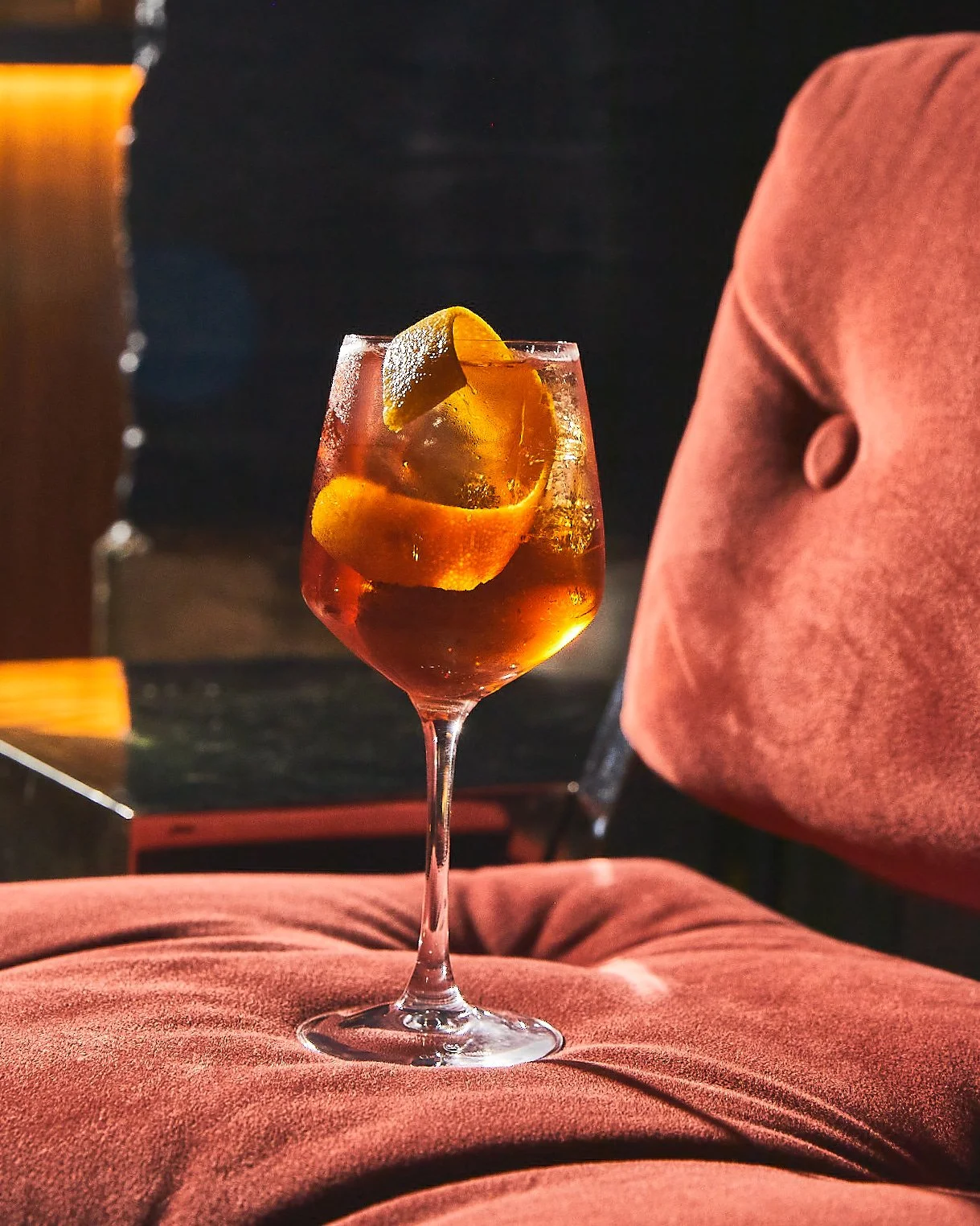 A cocktail in a stemmed glass with ice and lemon slice garnishing, resting on a red cushioned surface.