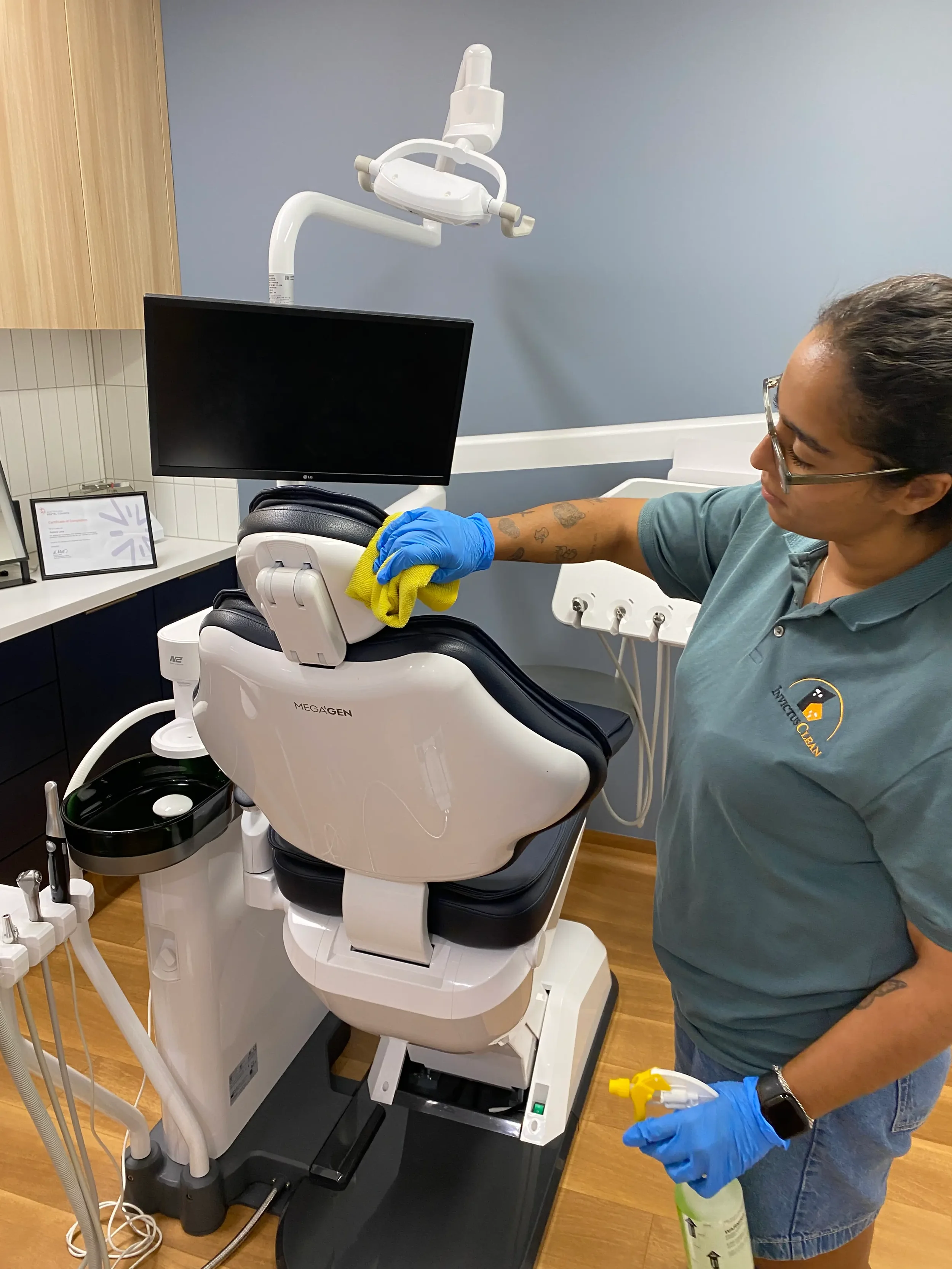 A dental hygienist in a gray scrub shirt and blue gloves cleaning a dental chair in a clinic.