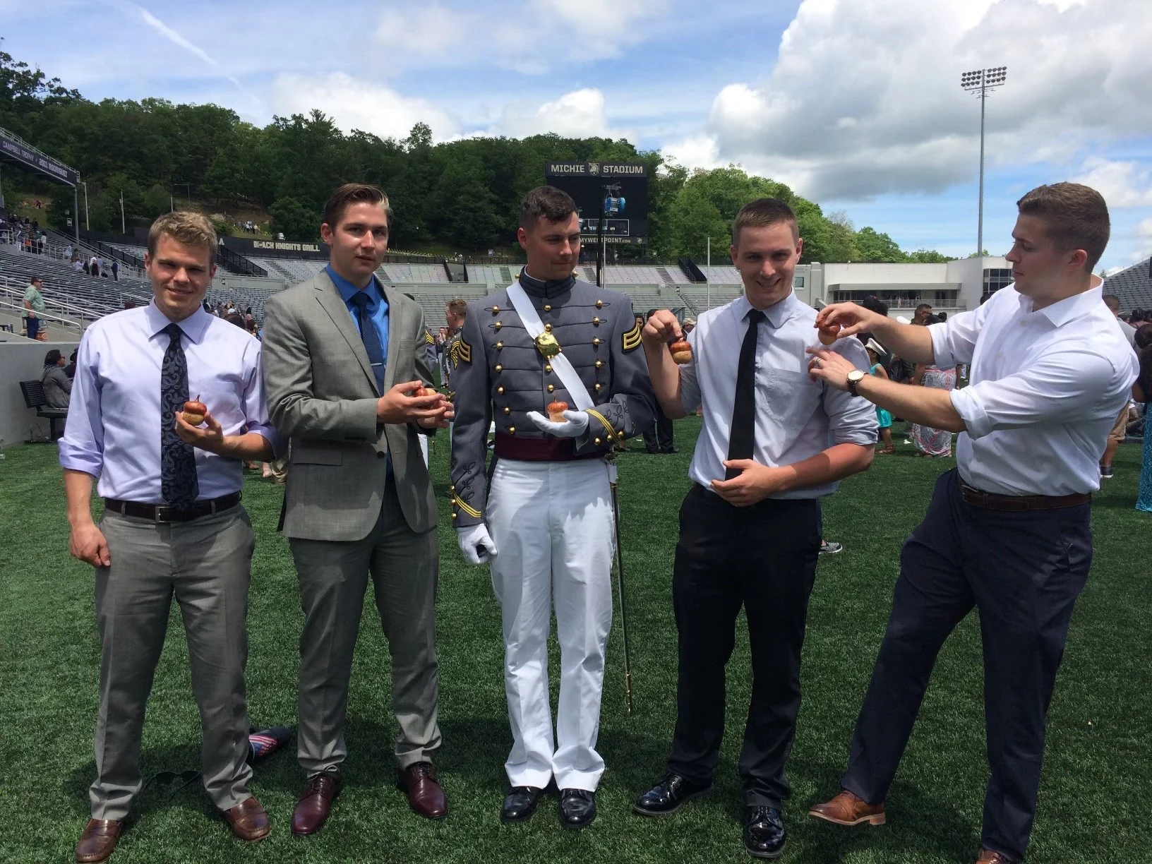 Five young men in formal and military attire standing on a sports field, holding muffins, with a stadium and trees in the background.