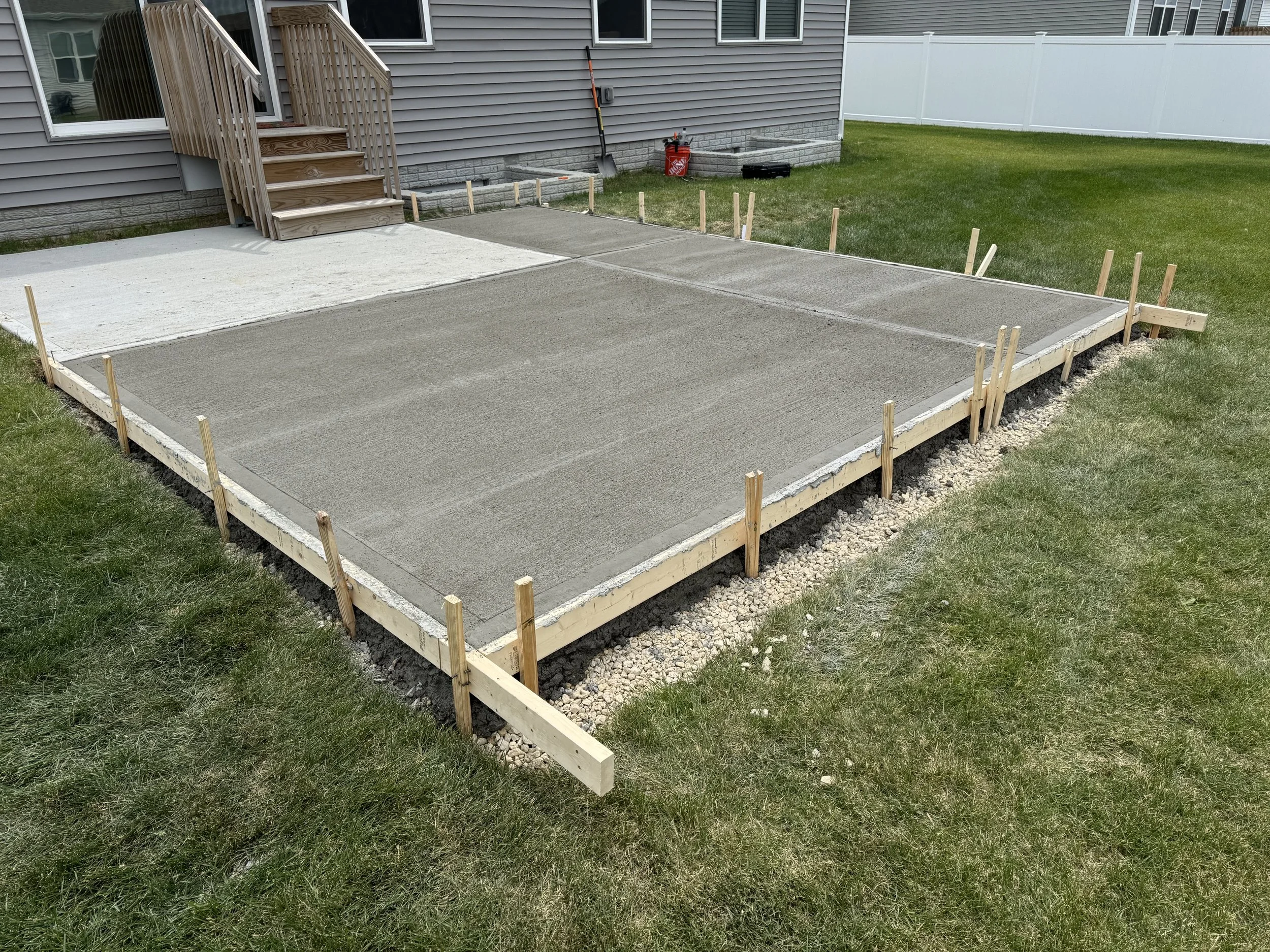 A backyard with a new concrete slab foundation for a building, surrounded by wooden framing and stakes, next to a house with a small wooden staircase.
