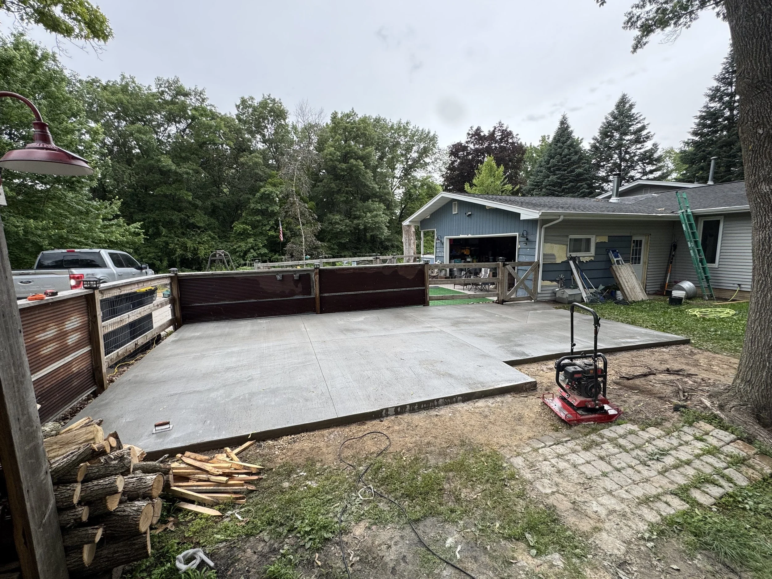 Backyard with newly poured concrete patio, construction tools, wood logs, a blue house with an attached garage, and green trees in the background.