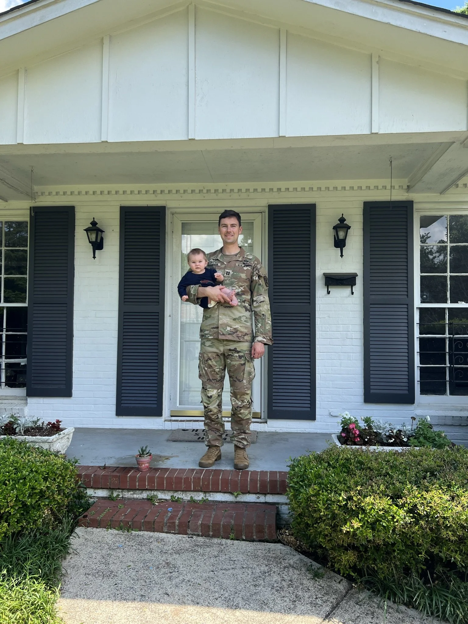 A man in military uniform holding a young child on his arm standing on a front porch of a white house with black shutters, plants, and flowers.