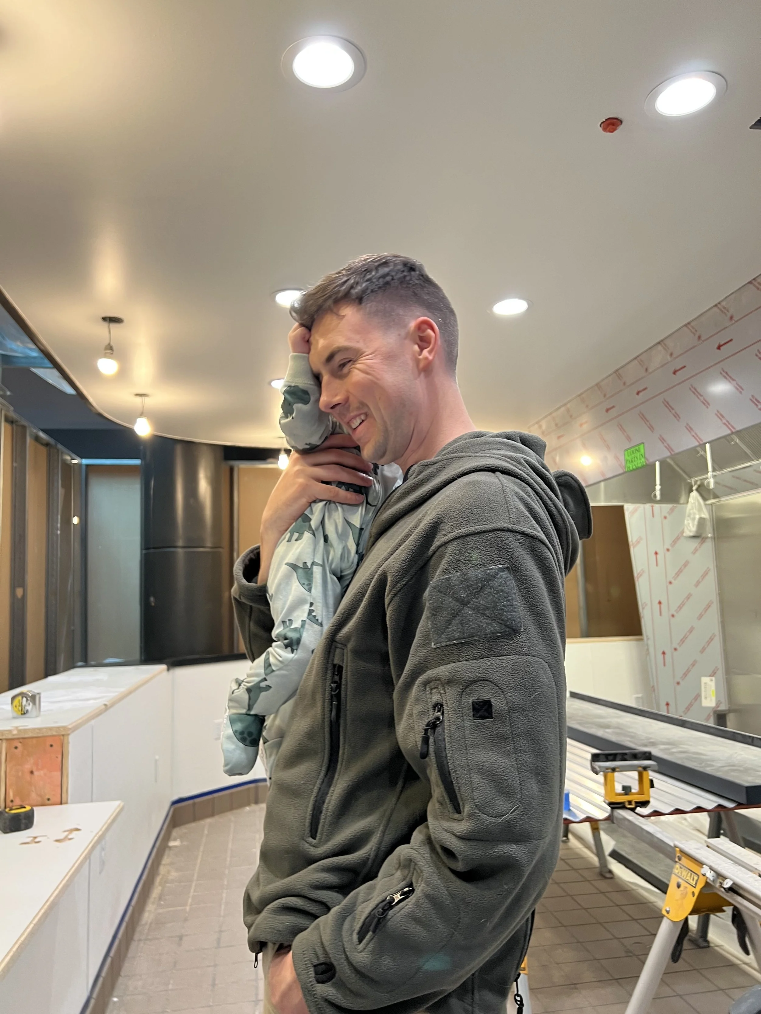 A man smiling and holding a child on his shoulder in a construction or renovation site indoors.
