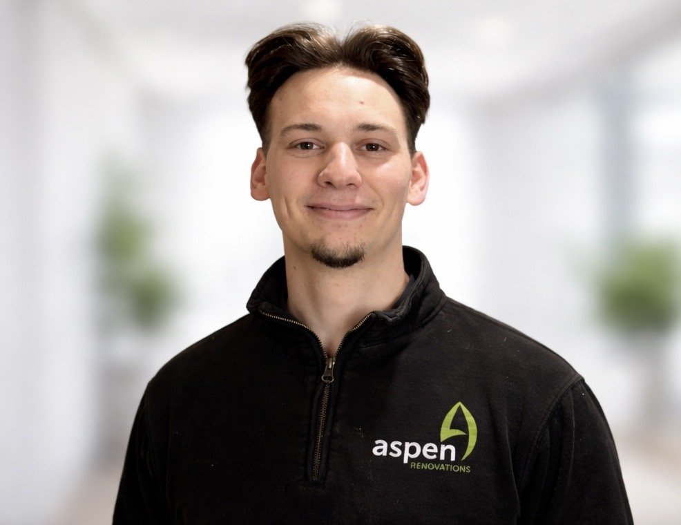 A young man with short, dark hair smiling while standing outside in front of a wooden fence. He is wearing a Black T-shirt with a logo that says 'aspen' on it. The sky is blue with a few clouds and there are houses with rooftops visible in the background.