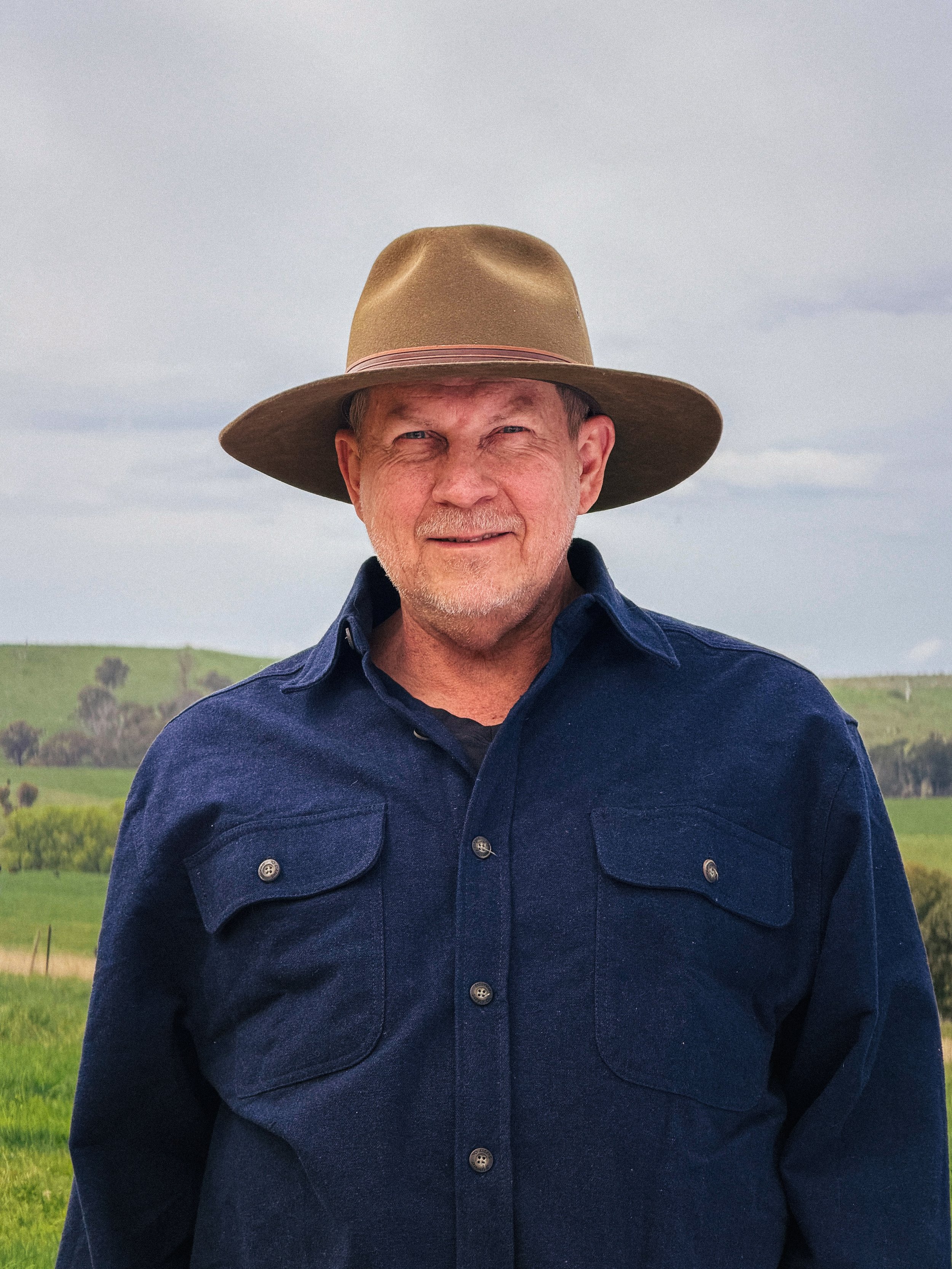 Older man wearing a wide-brimmed hat and a navy blue button-up shirt standing outdoors with green hills and cloudy sky in the background.