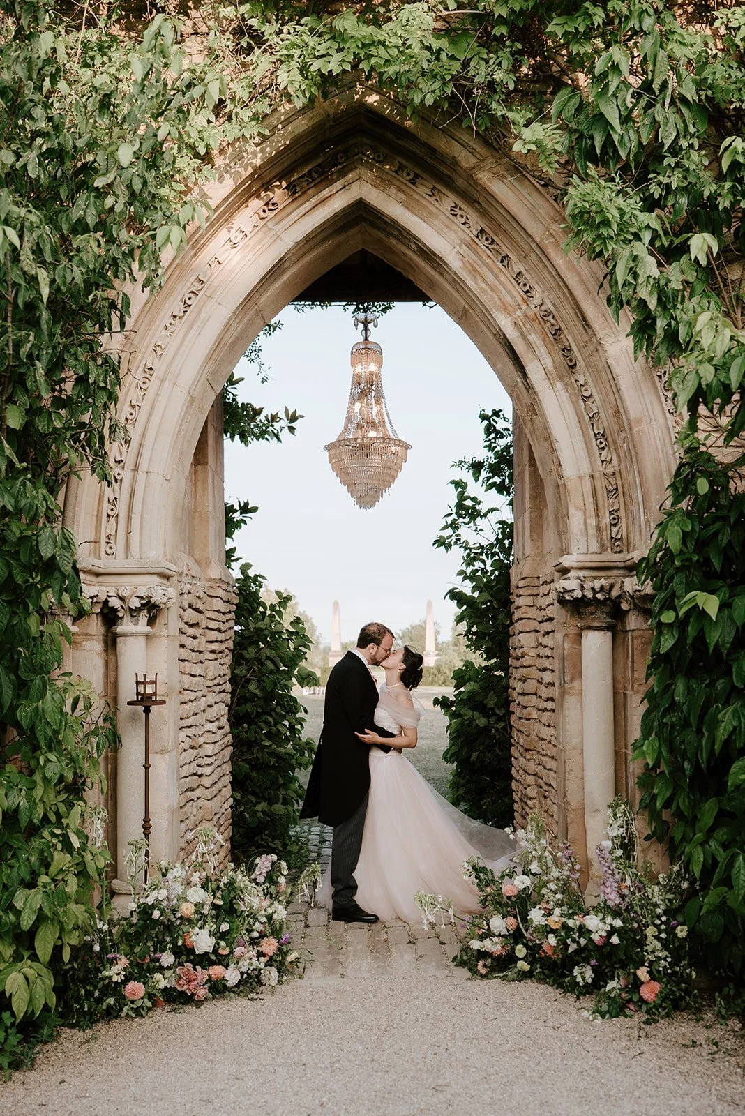 A bride and groom sharing a kiss under an ornate stone archway decorated with greenery, with a chandelier hanging overhead and a floral arrangement on either side of the arch.