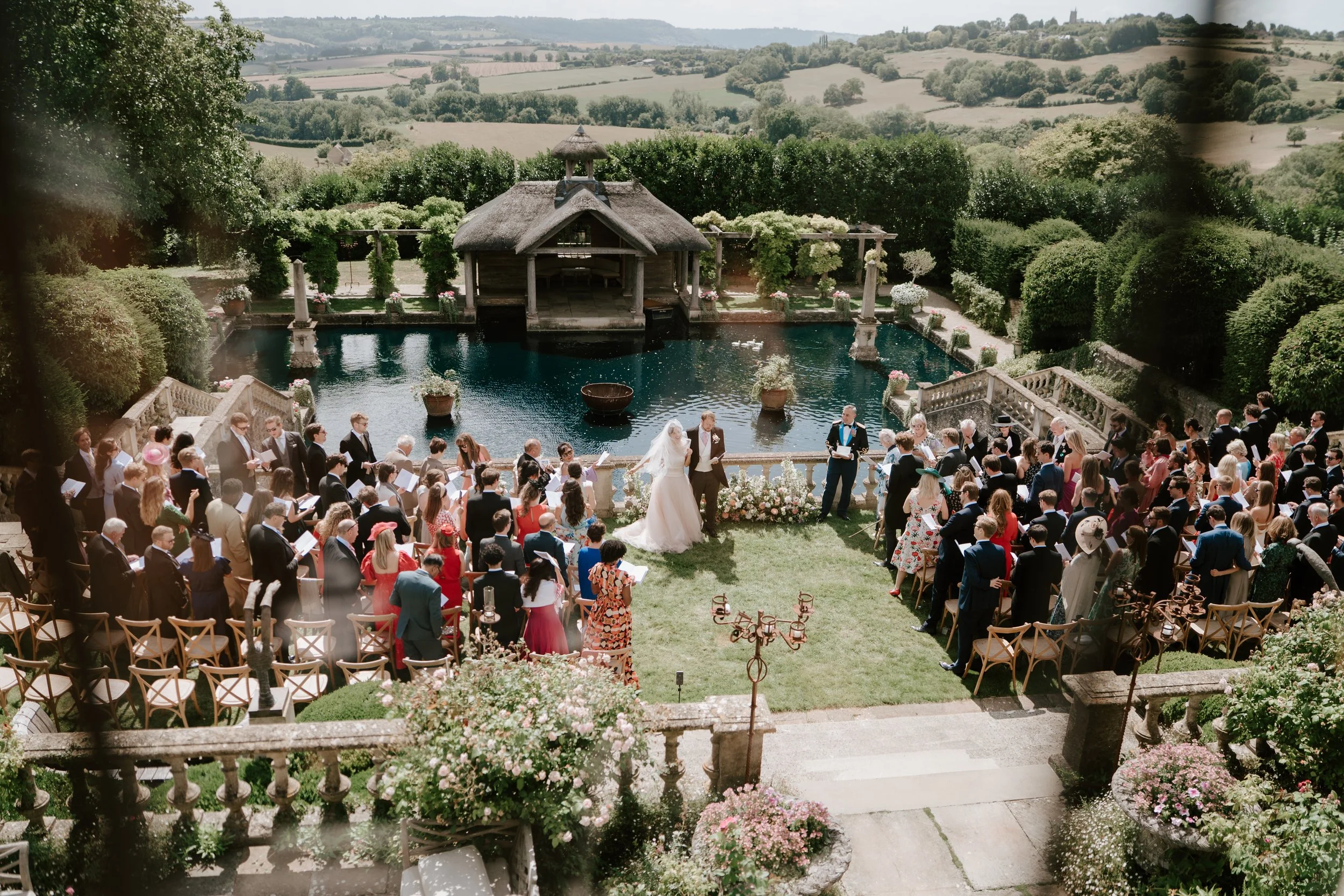 A wedding ceremony taking place outdoors by a pond with a thatched roof shelter in the background. The bride and groom are standing at the altar with guests seated and standing around them, facing the water. The setting is lush and decorated with flowers, surrounded by a scenic countryside landscape.