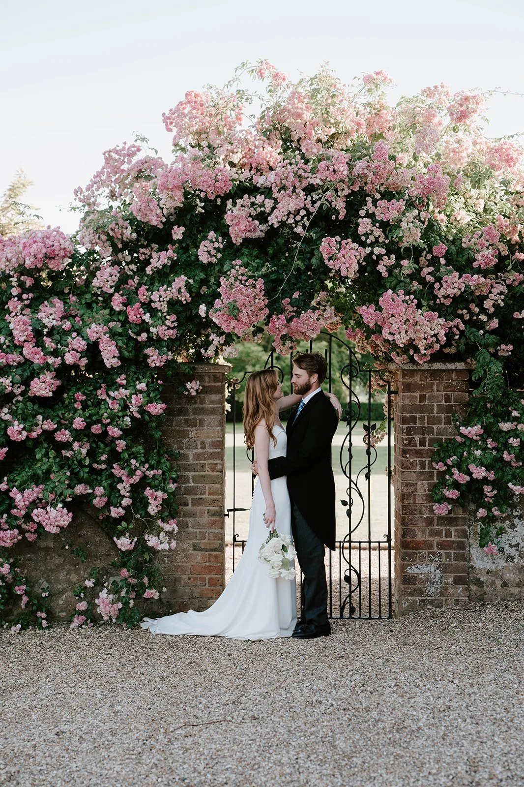 A bride and groom standing close together under a flowering pink tree arch and brick gate, holding hands, on their wedding day.