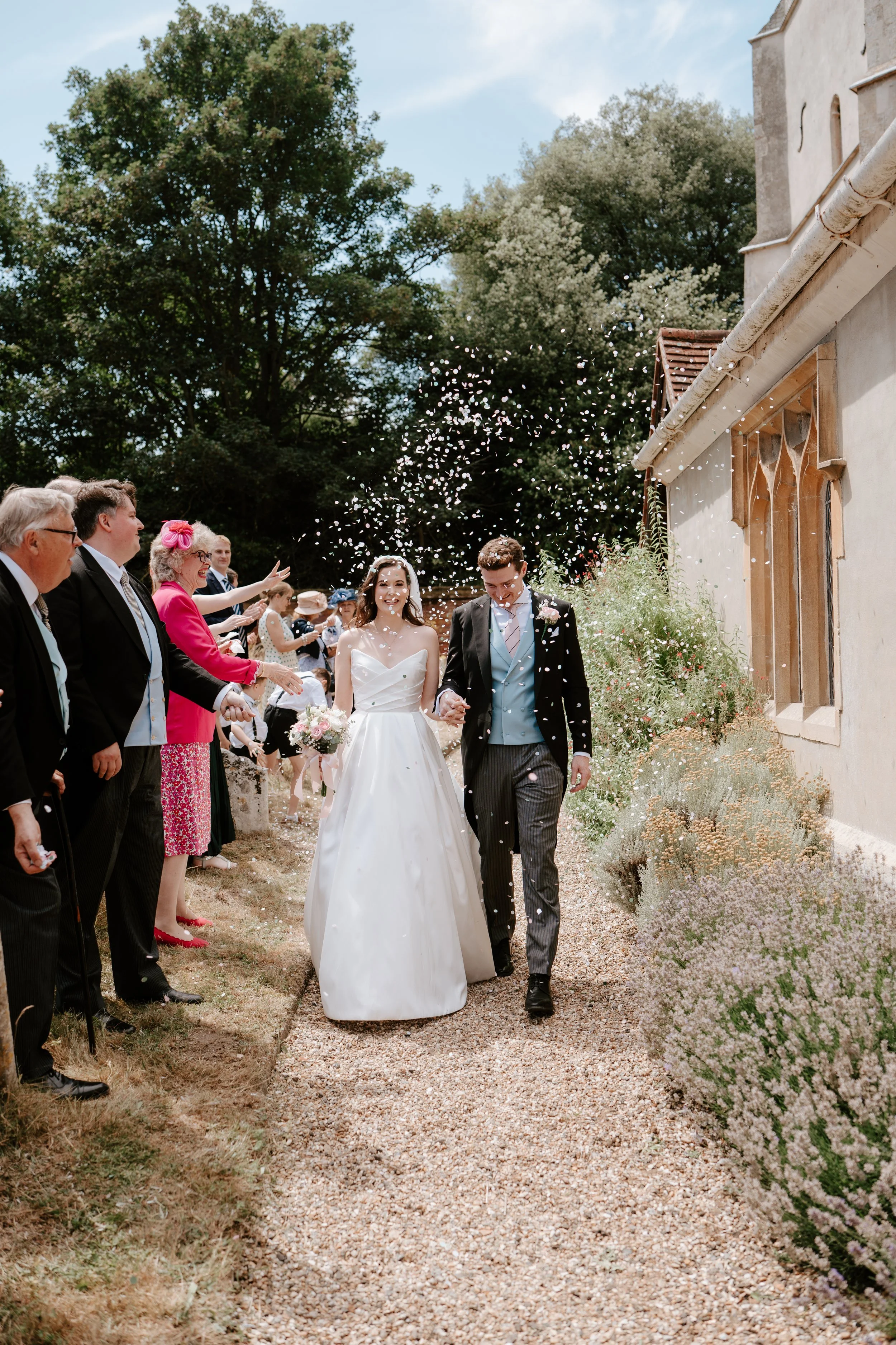 A bride and groom walking down an outdoor aisle at their wedding, surrounded by guests throwing confetti. The bride is in a white strapless wedding gown holding a bouquet, and the groom is in a dark suit with a vest and boutonniere. Guests are dressed formally, standing on either side of a gravel path with greenery and flowers on the side. A building with a tan wall and window is visible on the right side.