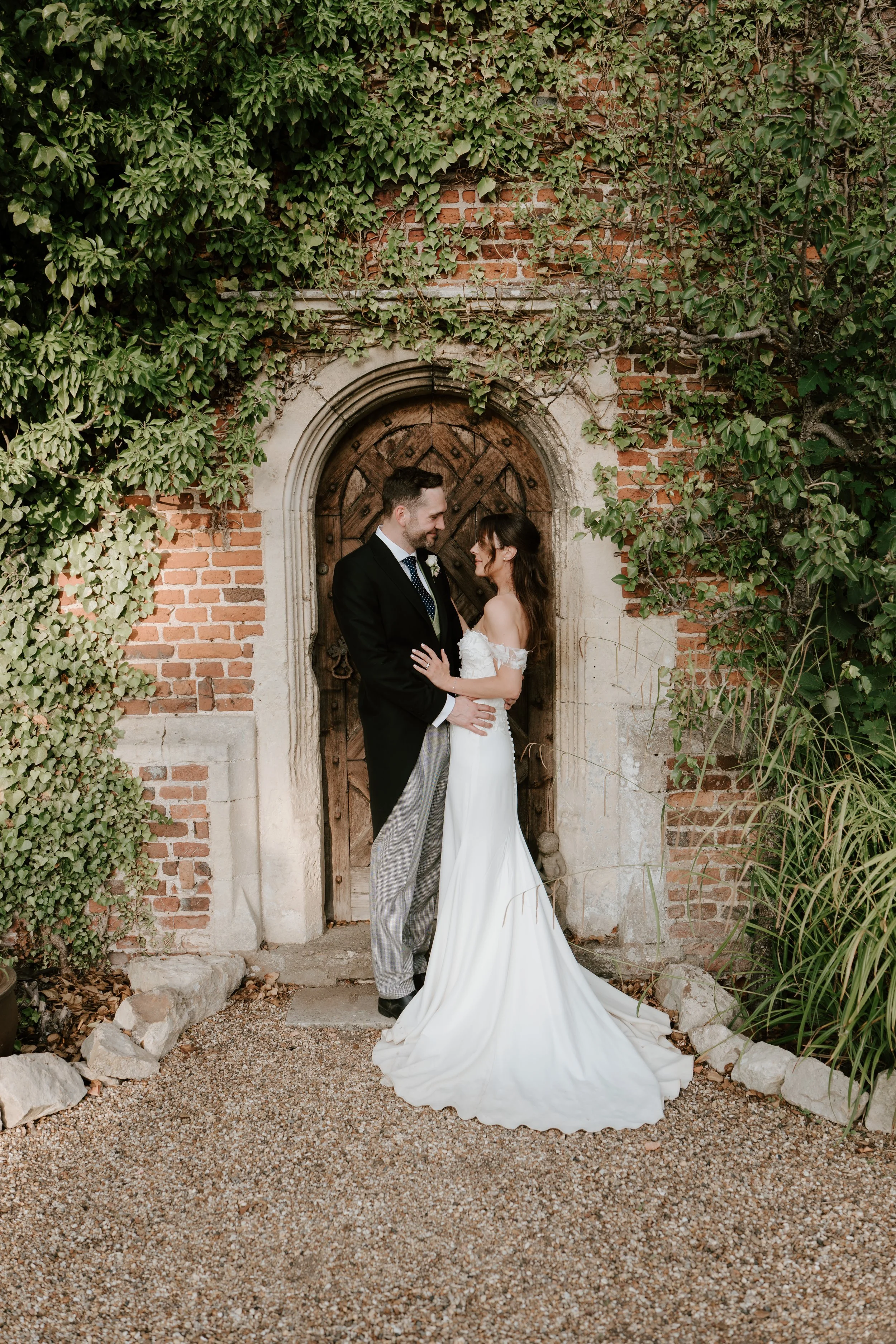 A bride and groom stand close together in front of a rustic wooden door surrounded by ivy and greenery, sharing a tender moment on their wedding day.