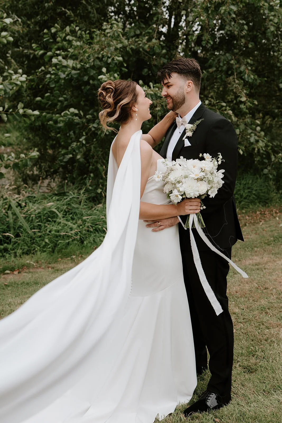 A bride and groom standing outdoors, smiling at each other, with the bride holding a bouquet of white flowers. The bride wears a white wedding gown with a flowing train, and the groom is dressed in a black tuxedo with a white bow tie. They are surrounded by green foliage.