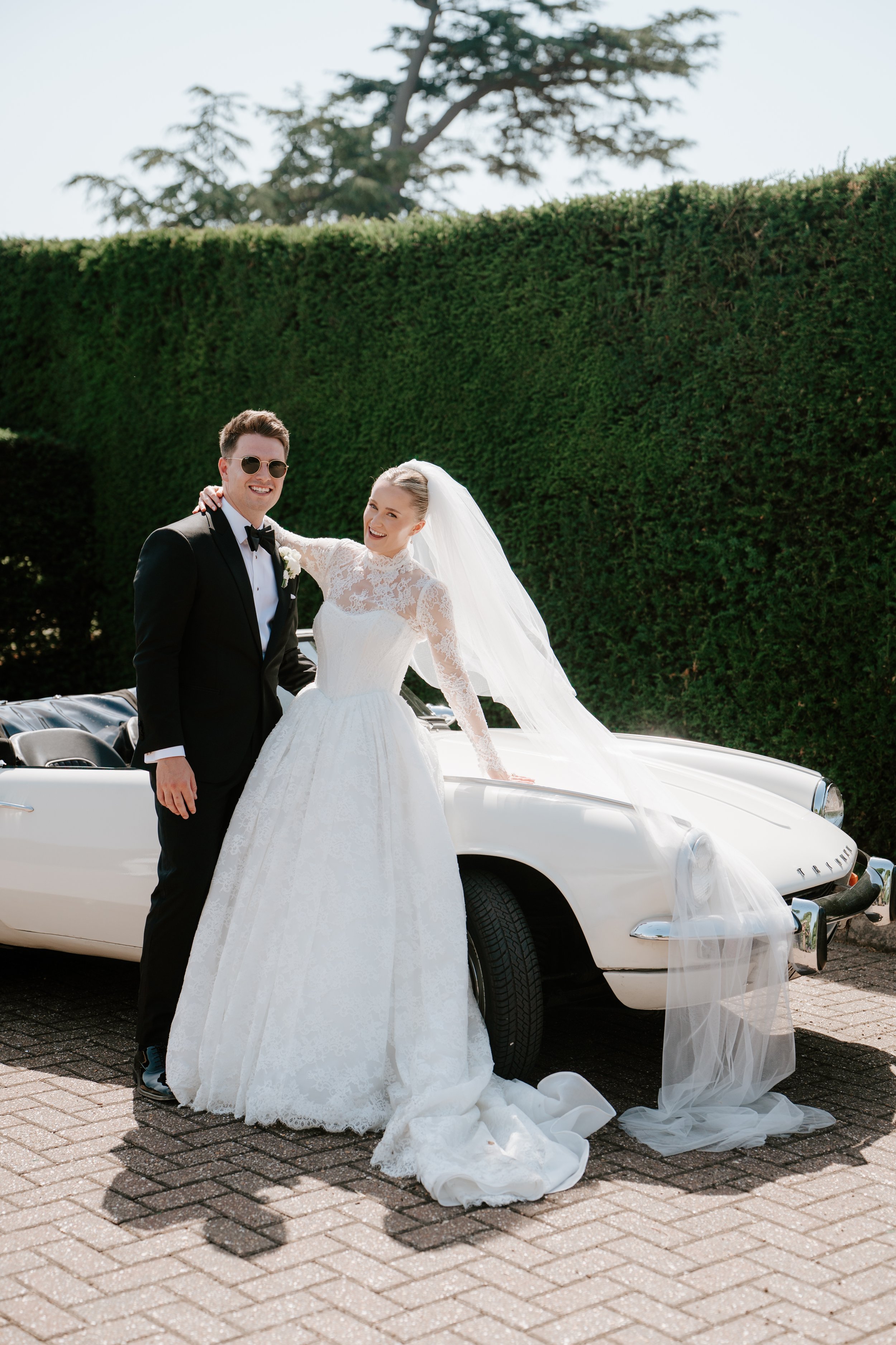 A bride and groom standing next to a vintage white car on a brick pavement. The bride is wearing a white lace wedding gown and veil, and the groom is in a black tuxedo with sunglasses. They are smiling, with the groom's arm around the bride's shoulder. There is green hedge and trees in the background.