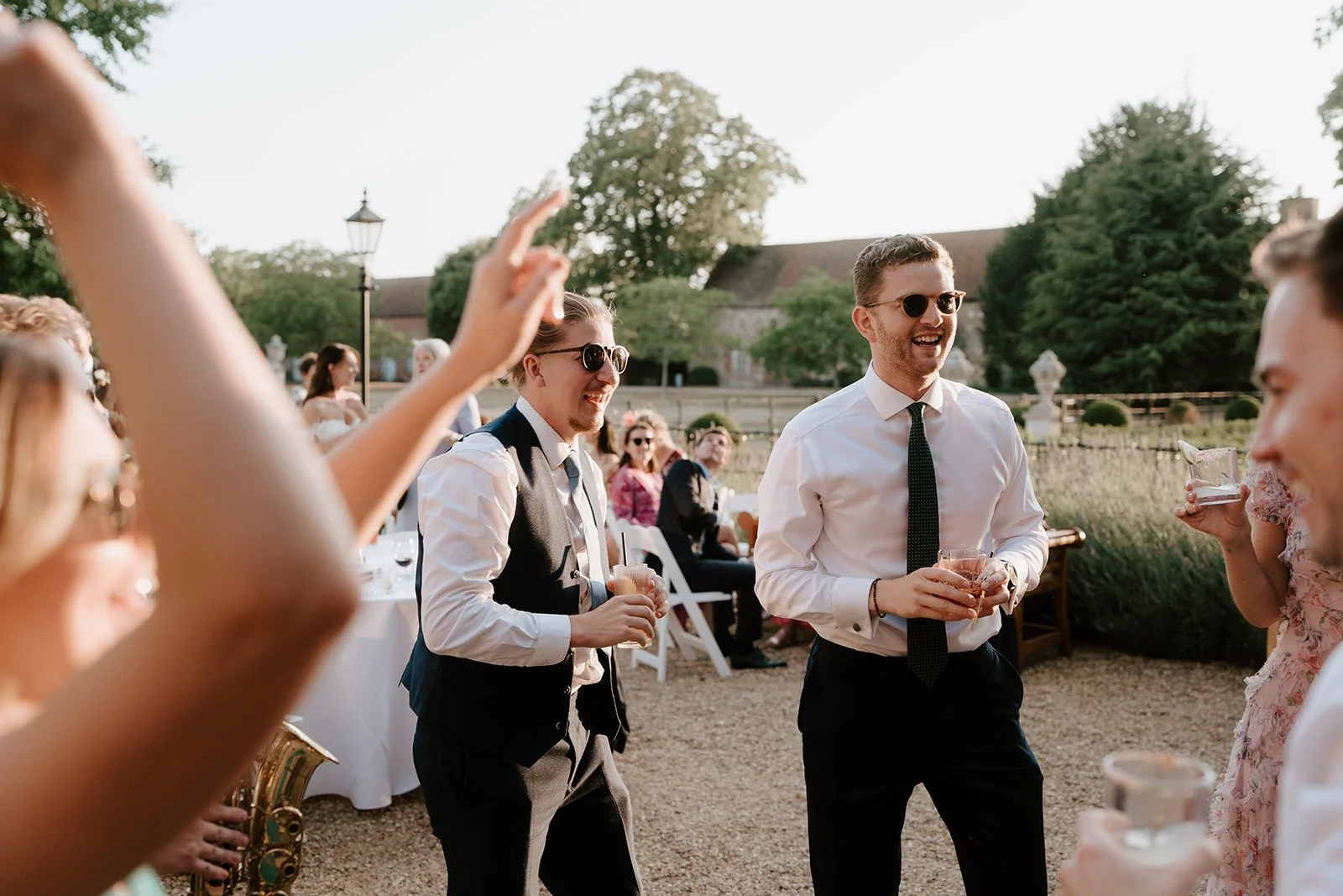 People at an outdoor party or wedding reception, smiling and holding drinks, with a garden and trees in the background.