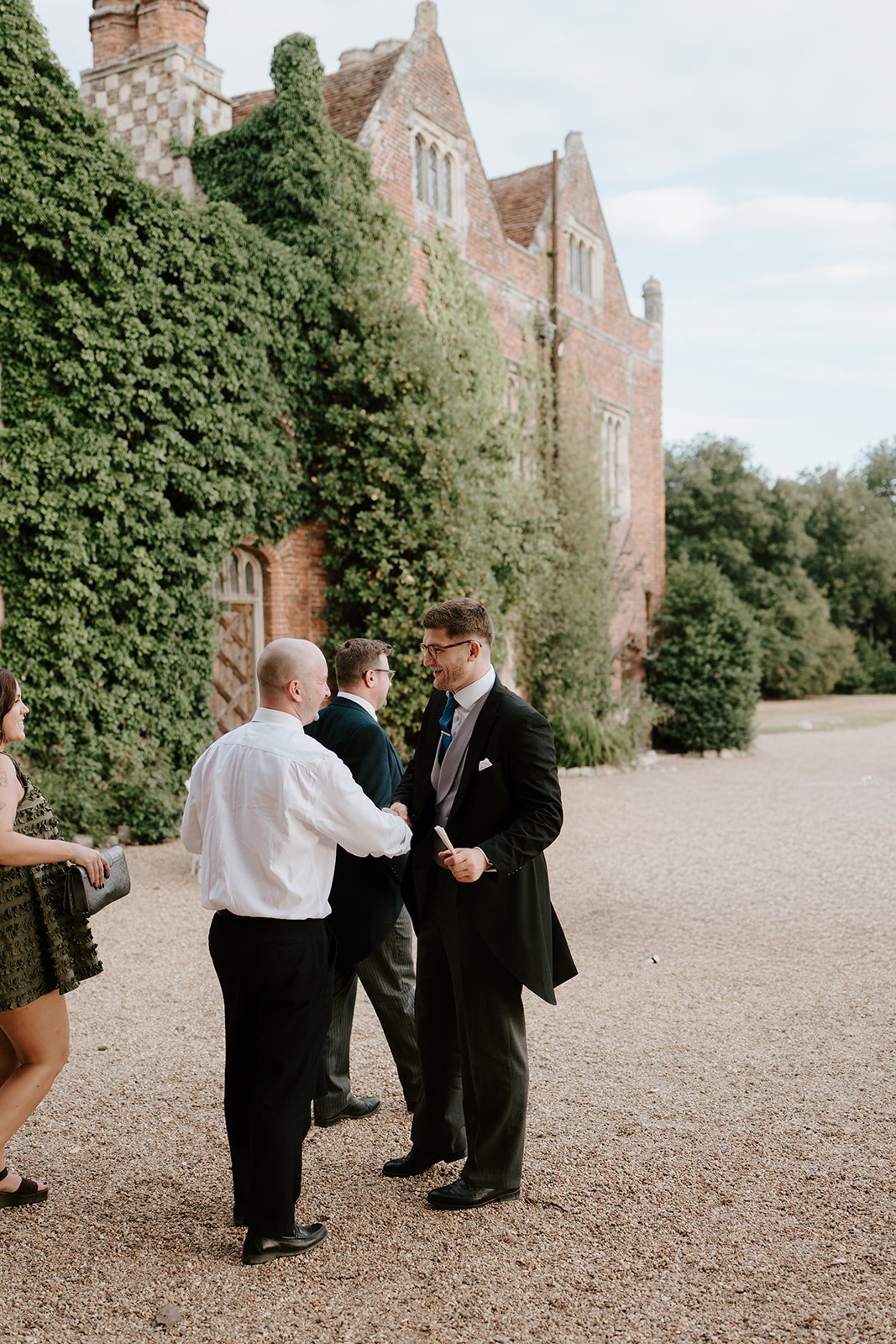 A group of people in formal attire smiling and shaking hands outside near an old brick building with green ivy. It appears to be a wedding or formal event.