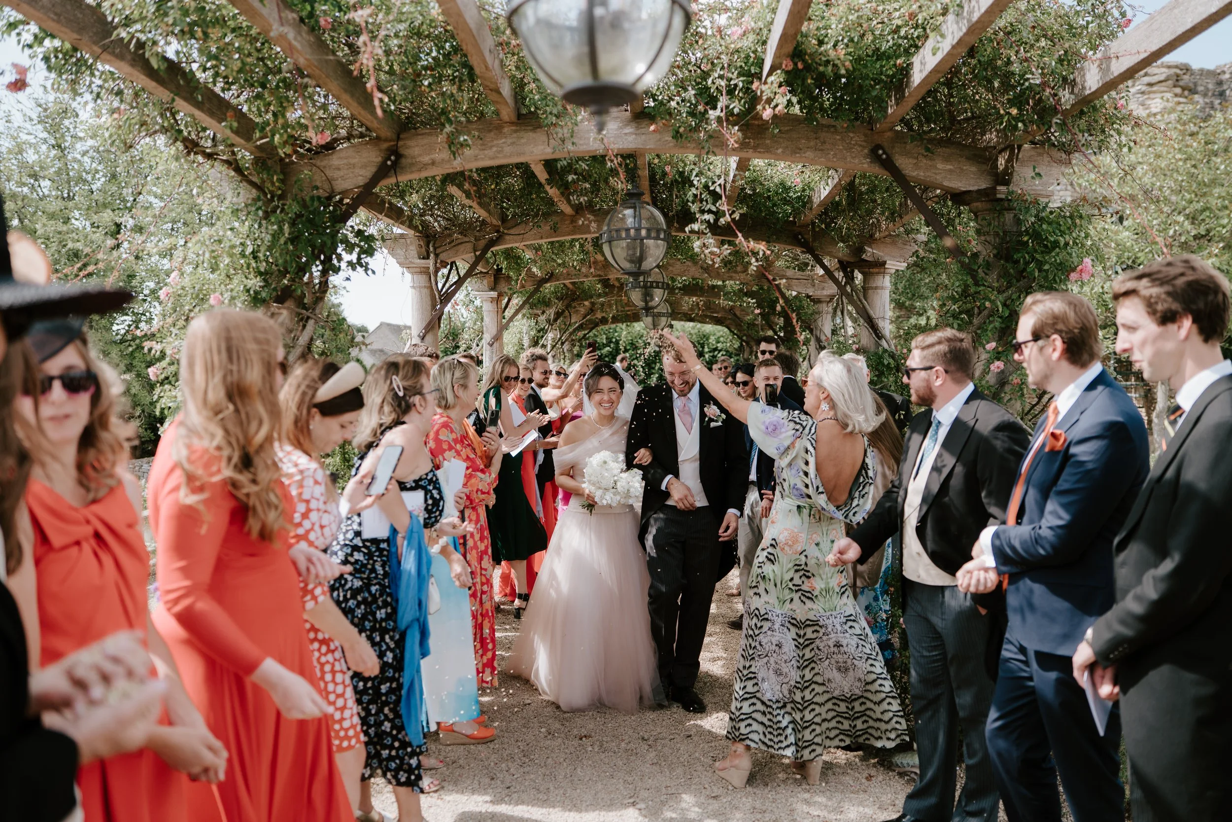 A bride and groom smiling and holding hands under a wooden trellis decorated with flowers, surrounded by wedding guests outdoors.