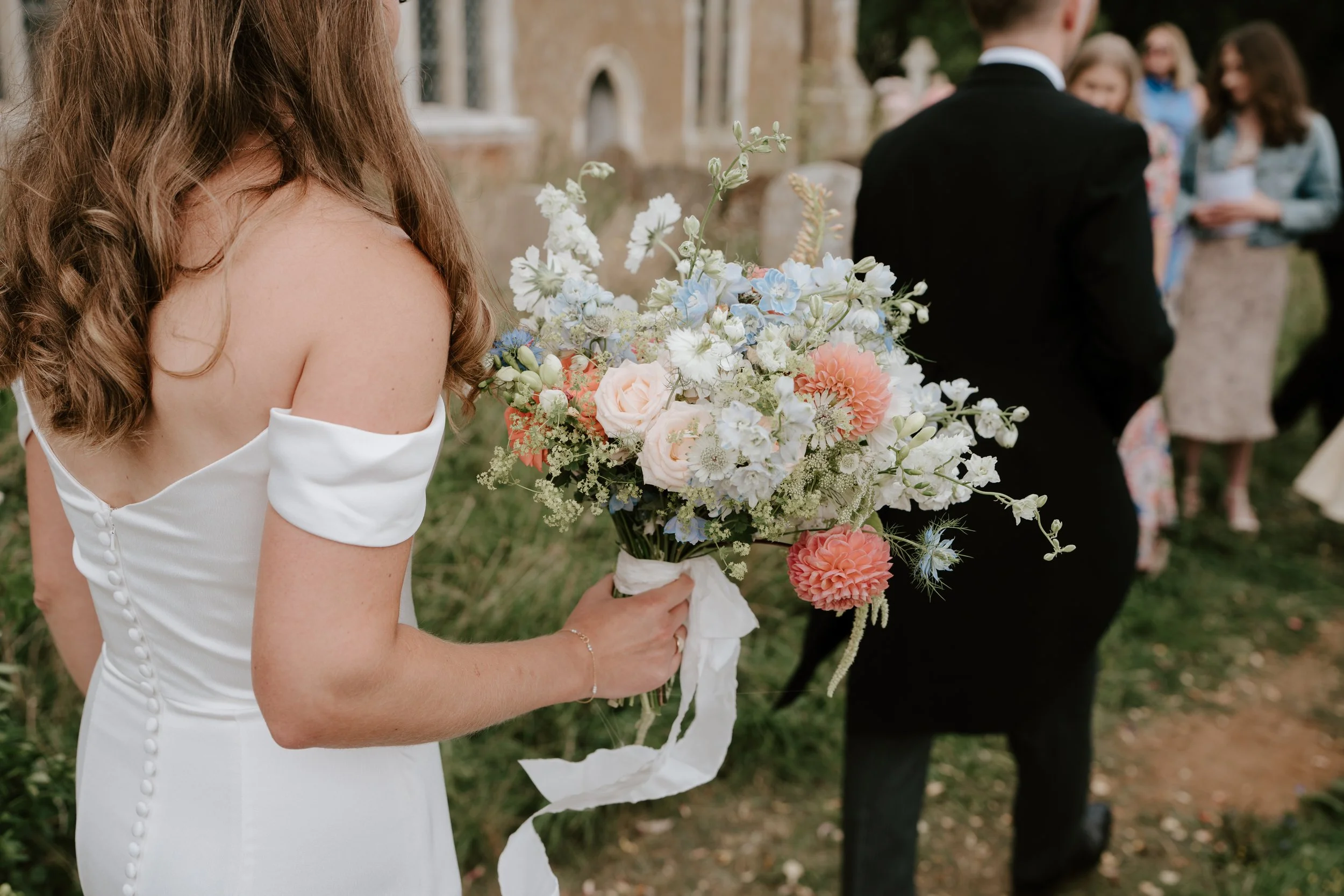 A woman in a white dress holding a bouquet of pink, white, and blue flowers during a wedding ceremony outdoors, with people in formal attire in the background.