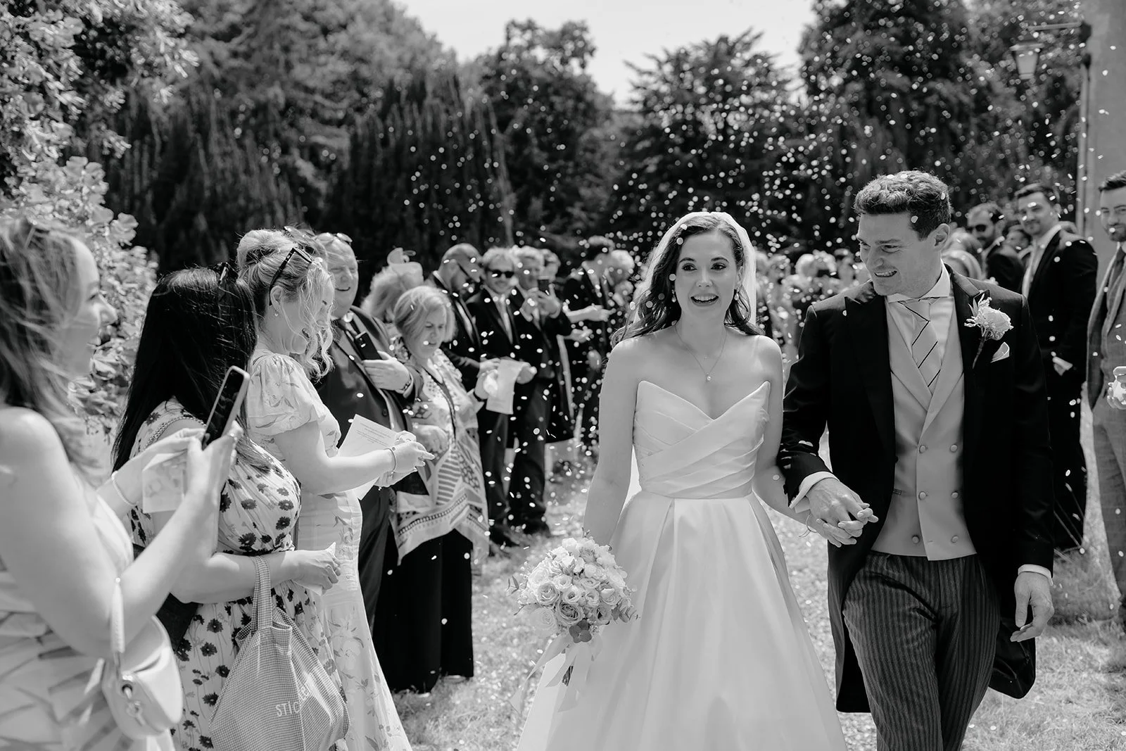 A black-and-white photo of a bride and groom walking outdoors after their wedding, holding hands, with guests celebrating and throwing confetti around them.
